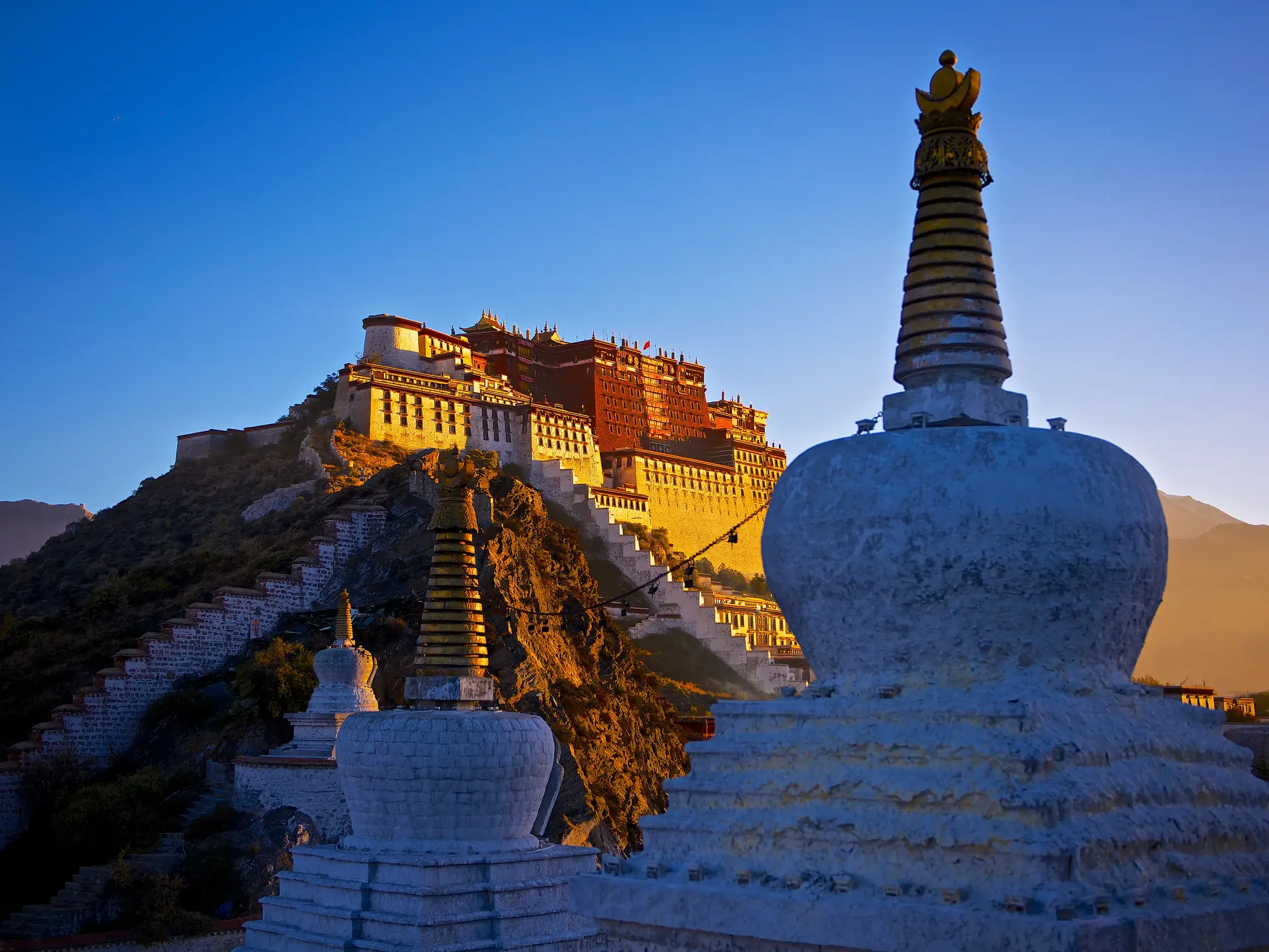 Der Potala-Palast in Lhasa thront im warmen Licht des Sonnenaufgangs über weißen buddhistischen Stupas im Vordergrund.