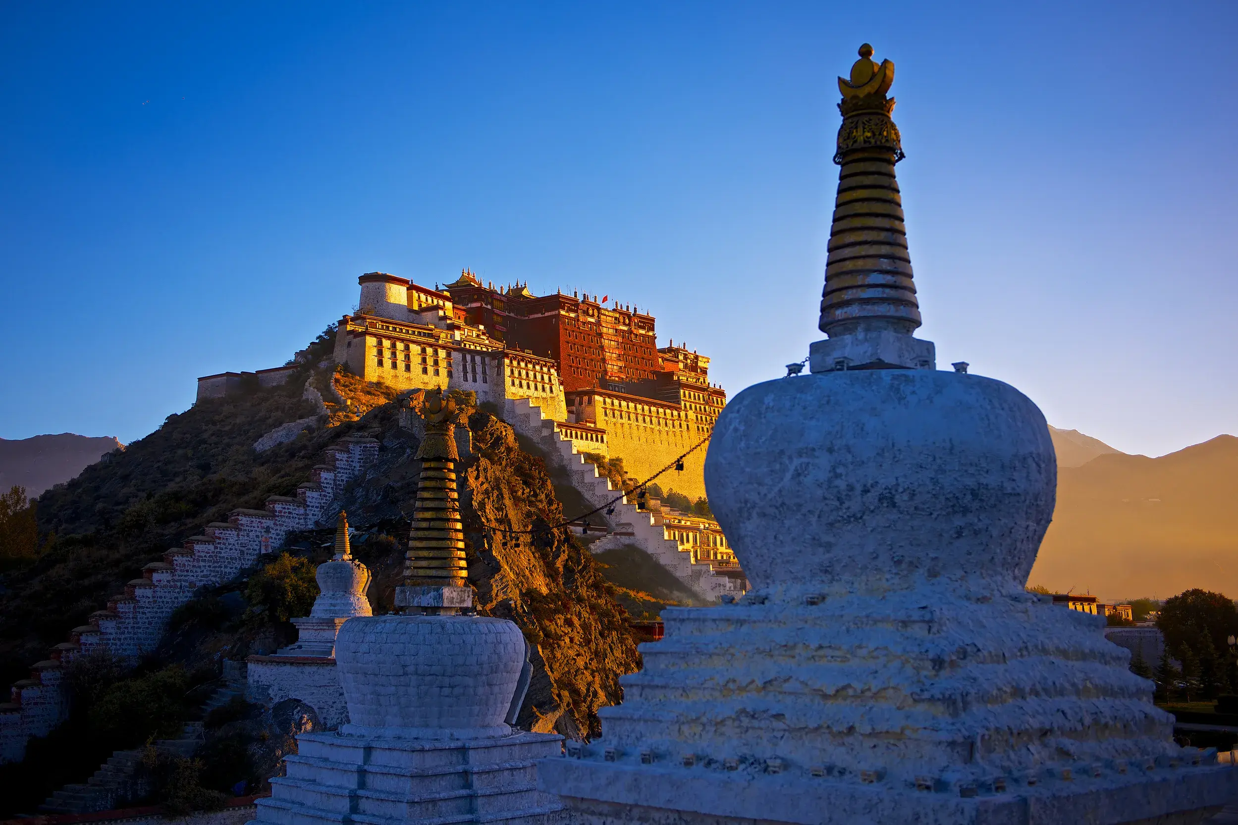 Der Potala-Palast in Lhasa thront im warmen Licht des Sonnenaufgangs über weißen buddhistischen Stupas im Vordergrund.