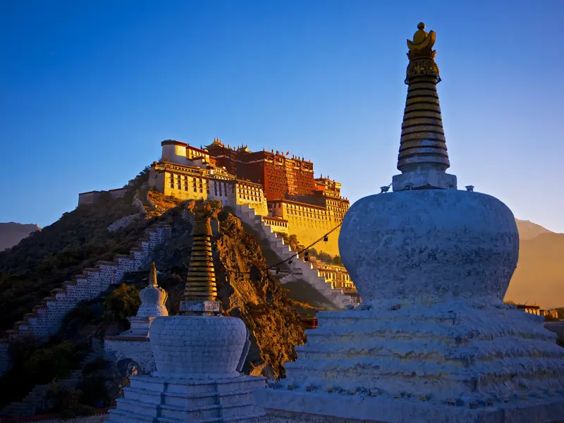 Der Potala-Palast in Lhasa thront im warmen Licht des Sonnenaufgangs über weißen buddhistischen Stupas im Vordergrund.