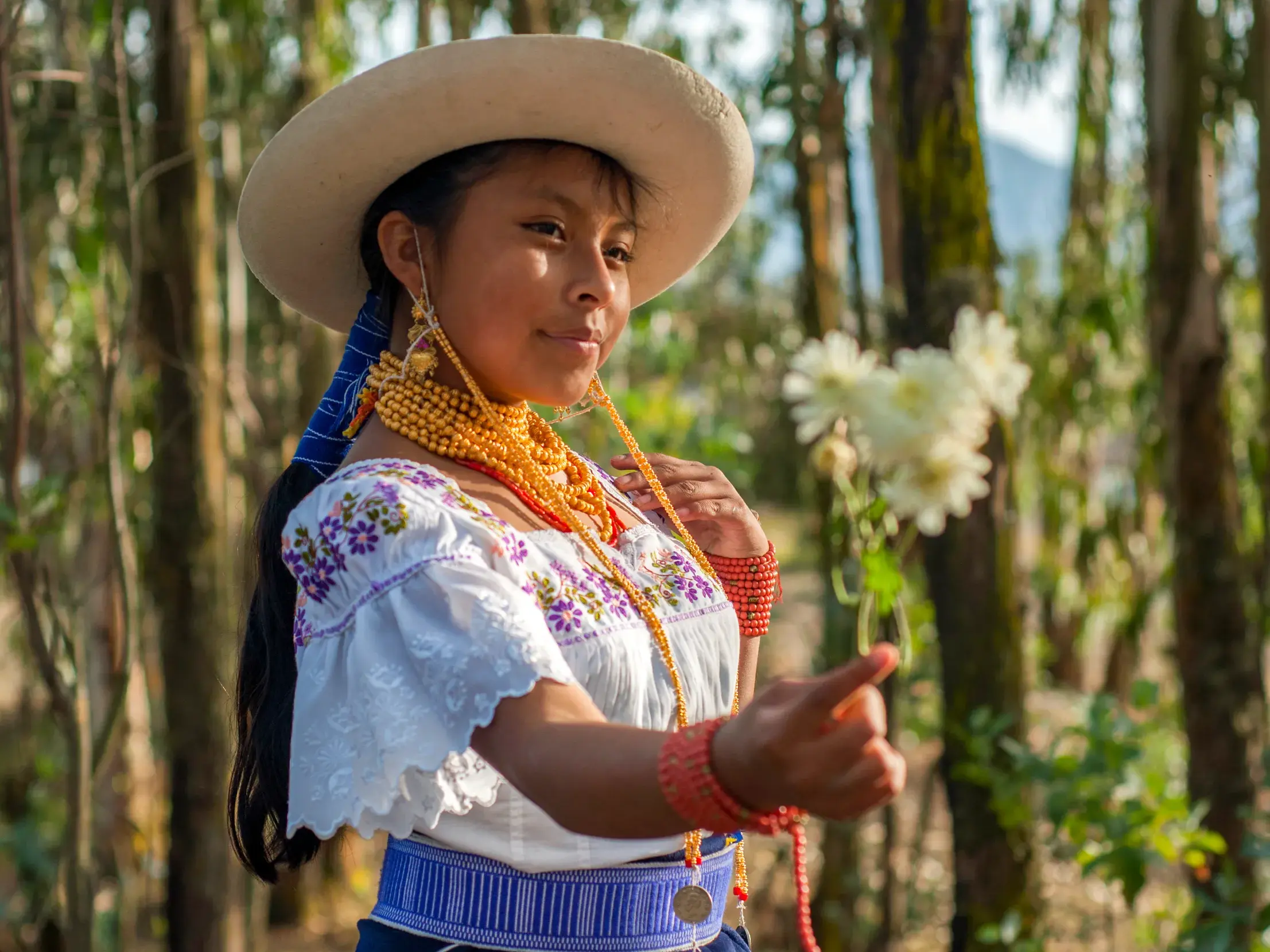 Porträt einer Frau in ecuadorianischer Tracht mit Hut und goldenem Halsschmuck vor Bäumen im Andenhochland.