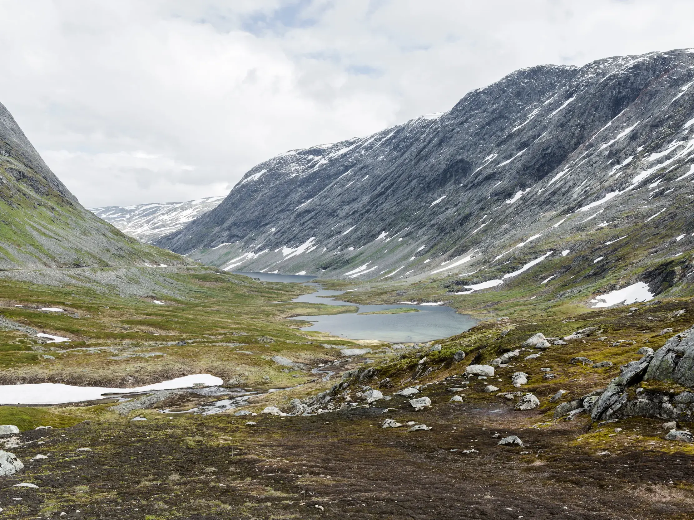Weite, karge Berglandschaft im Jotunheimen Nationalpark mit Schneefeldern und kleinen Seen im Tal unter bewölktem Himmel.