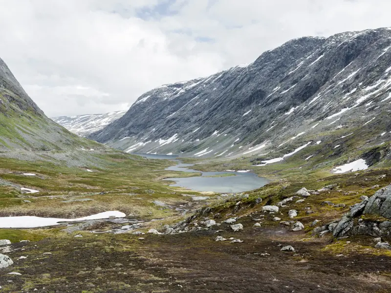 Weite, karge Berglandschaft im Jotunheimen Nationalpark mit Schneefeldern und kleinen Seen im Tal unter bewölktem Himmel.