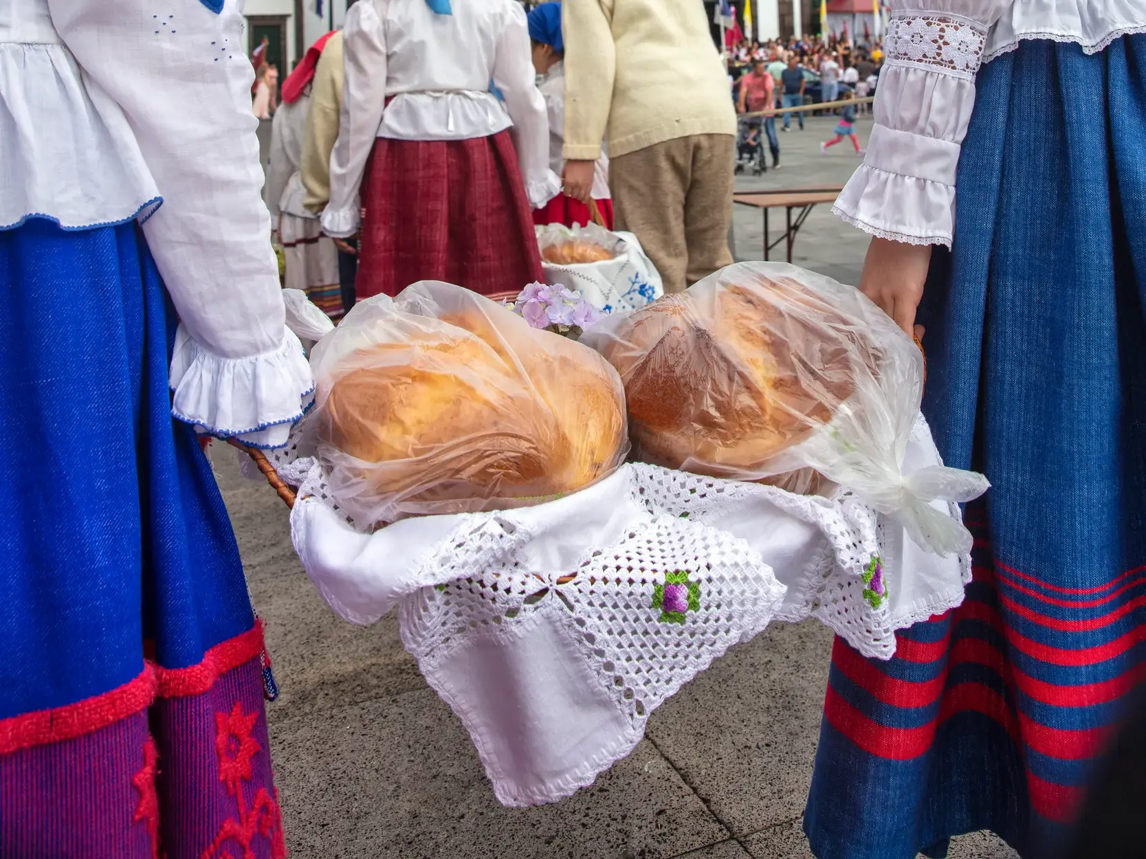 Frauen in farbenfrohen, traditionellen Trachten der Azoren präsentieren frisches, lokales Brot in einem Weidenkorb.