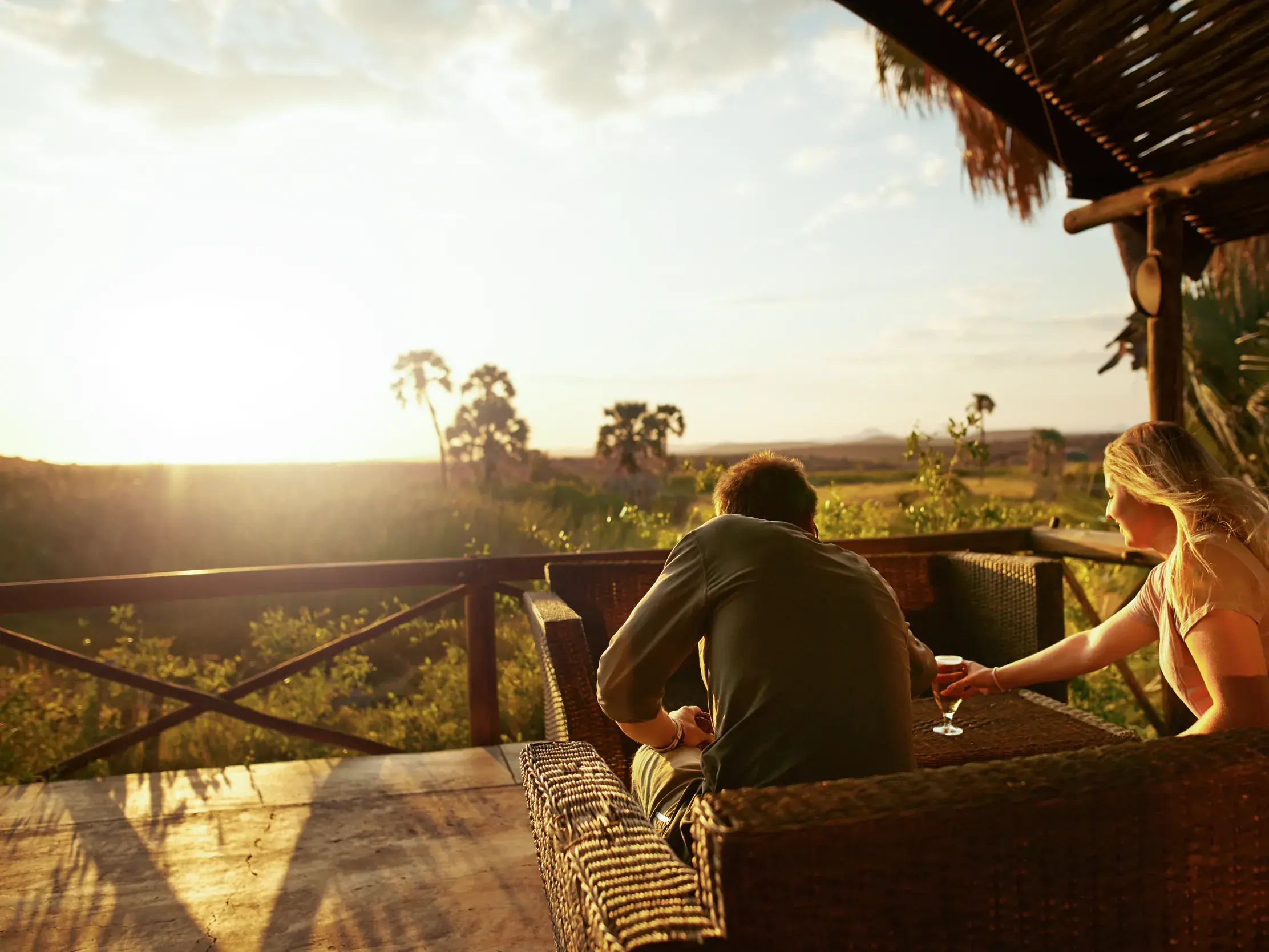 Ein Paar entspannt bei Sonnenuntergang mit einem Glas Wein auf der Holzveranda einer Lodge mit Blick auf die Savanne Namibias.