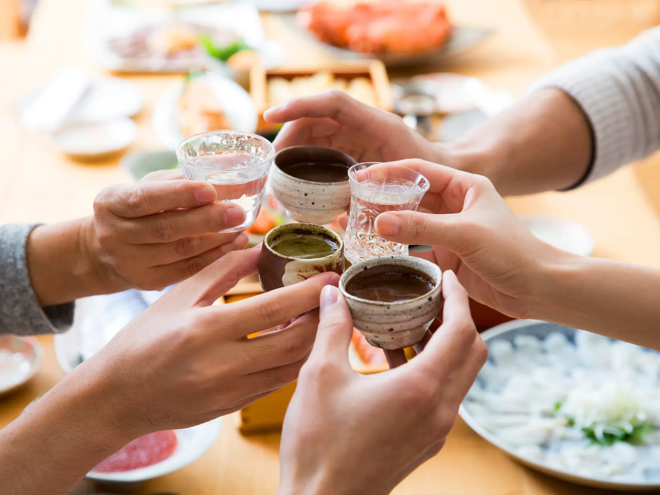 Hände einer Reisegruppe stoßen bei einer Verkostung in Japan mit traditionellen Sake- und Teeschalen an.