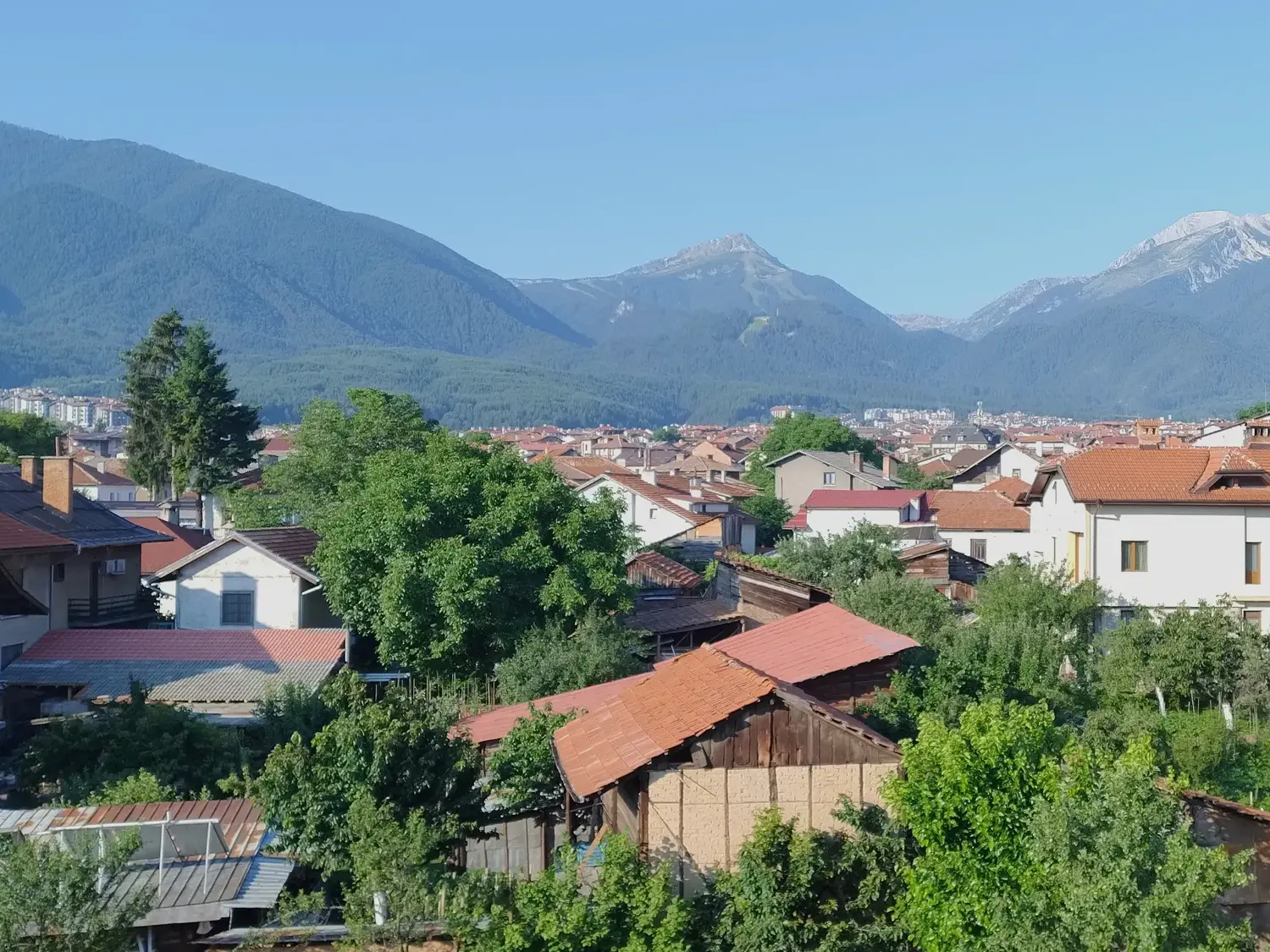 Sonniger Ausblick über die roten Dächer von Bansko auf die bewaldeten Hänge und felsigen Gipfel des Pirin-Gebirges in Bulgarien.