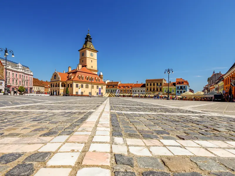 Tiefer Blick über das Kopfsteinpflaster des Rathausplatzes in Kronstadt auf das historische Rathaus unter blauem Himmel.