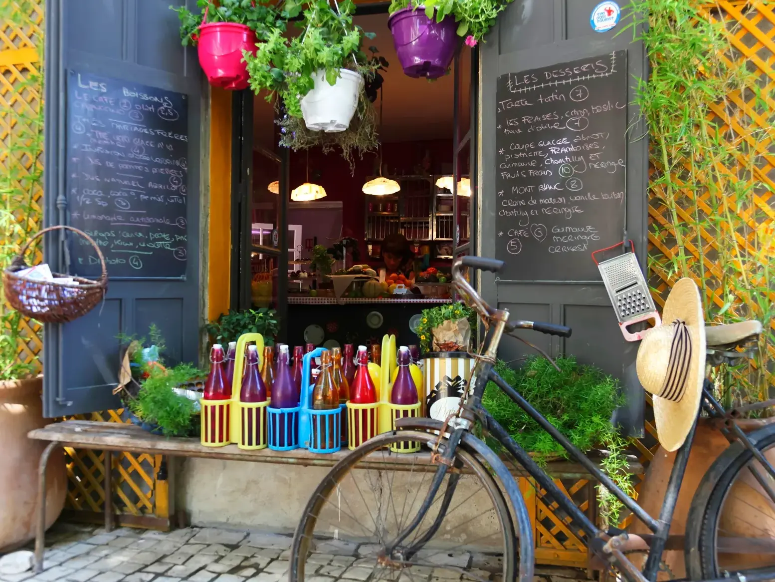 Ein Fahrrad mit Strohhut parkt vor einem charmanten Café in der Provence mit einer Menü-Tafel und bunten Limonaden.