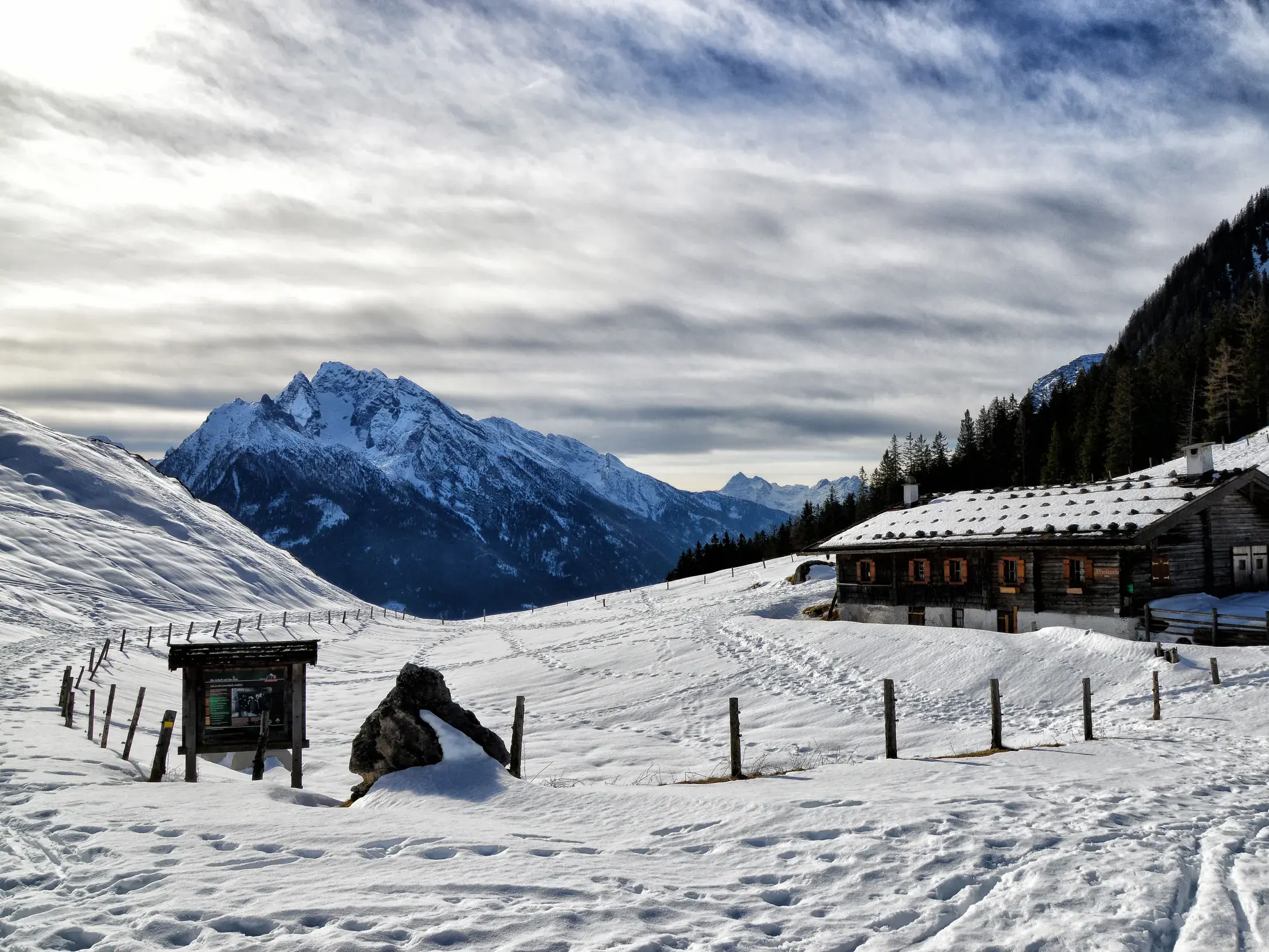Rustikale Almhütte in weiter Schneelandschaft mit Spuren im Schnee und Blick auf die Gipfel der Berchtesgadener Alpen.