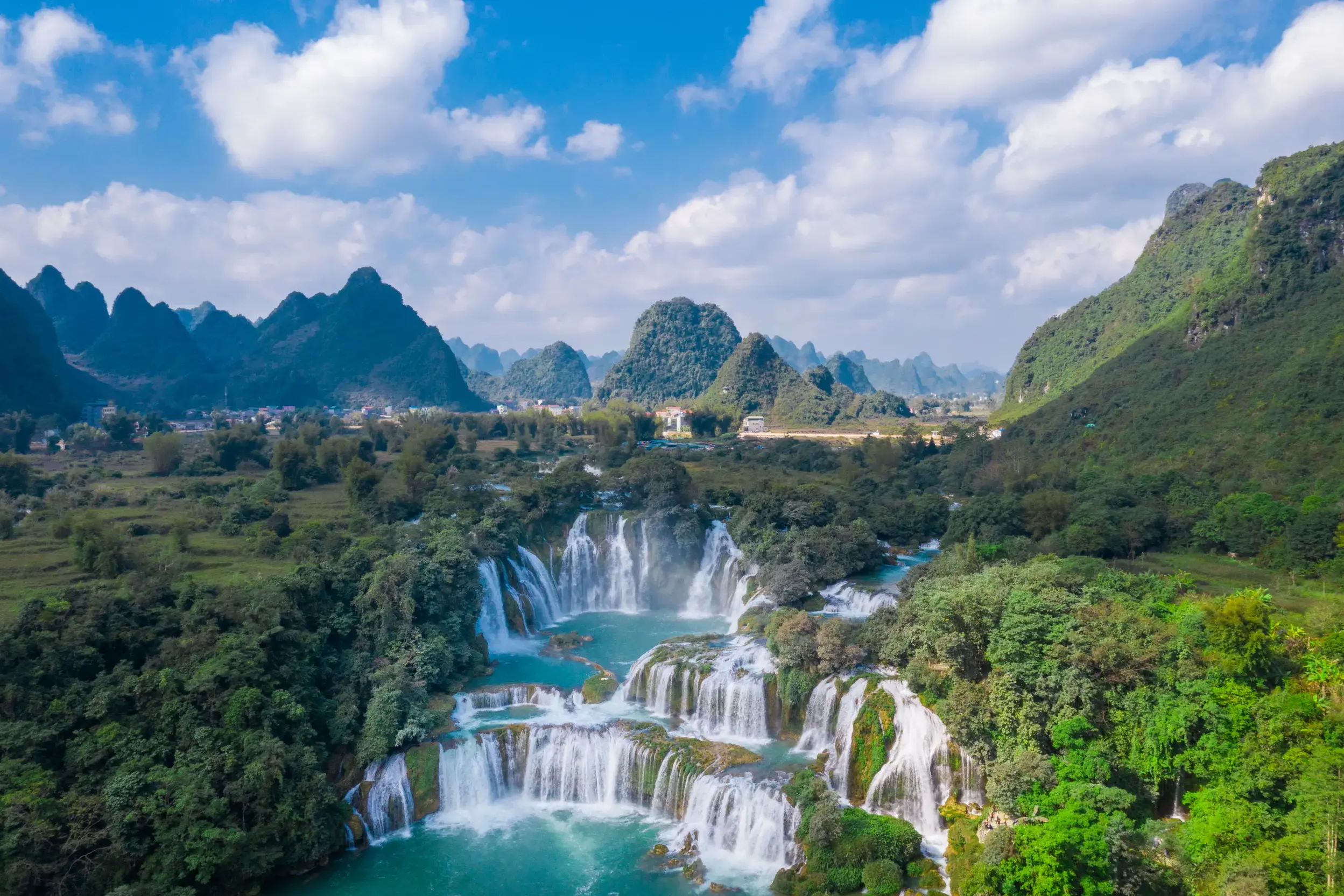 Luftaufnahme der mehrstufigen Ban-Gioc-Wasserfälle in Nordvietnam, umgeben von grünen Karstbergen unter blauem Himmel.