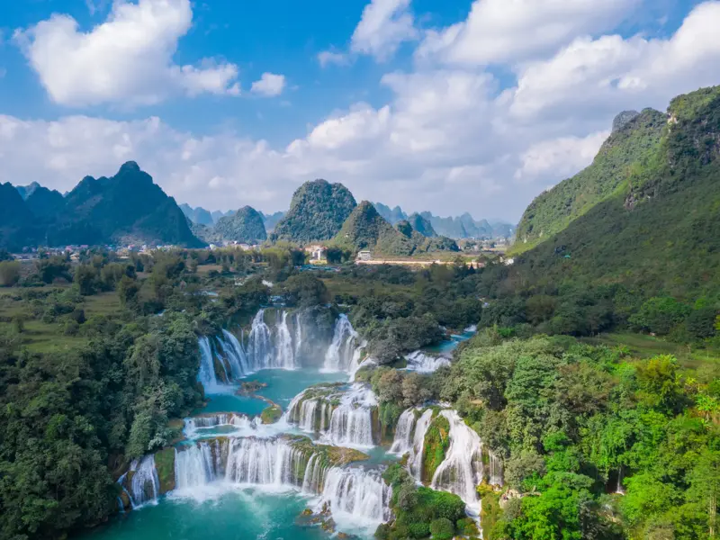 Luftaufnahme der mehrstufigen Ban-Gioc-Wasserfälle in Nordvietnam, umgeben von grünen Karstbergen unter blauem Himmel.