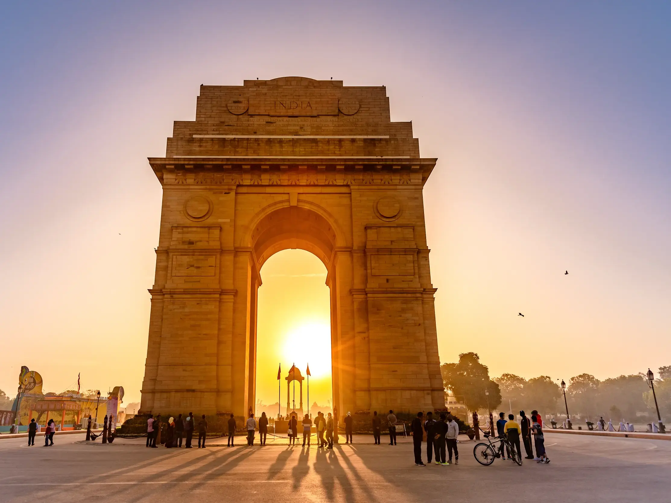Eine Gruppe junger Leute auf einer Rundreise steht vor dem India Gate in Delhi, das im goldenen Sonnenaufgang erstrahlt.