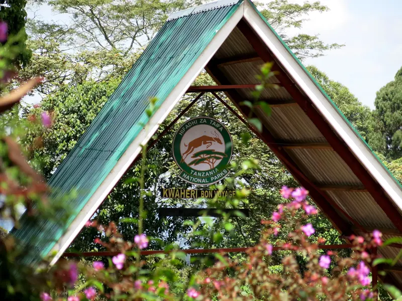 Abschied vom Kilimanjaro: Das "Kwaheri"-Schild am Marangu Gate des Nationalparks umrahmt von tropischer Vegetation und Blüten.