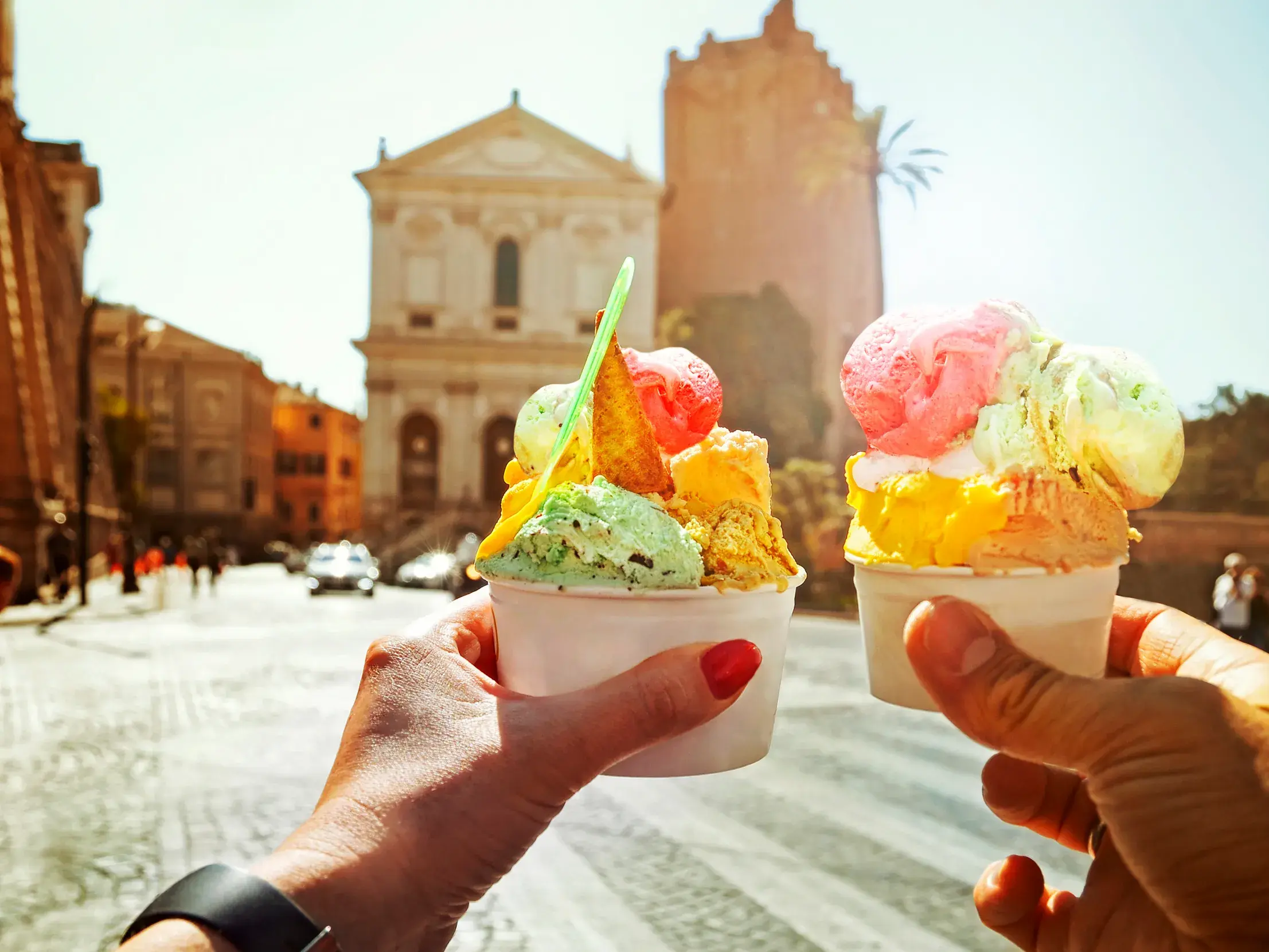 Zwei Hände halten Becher mit buntem Gelato vor einer historischen Fassade in den sonnigen Gassen von Rom.