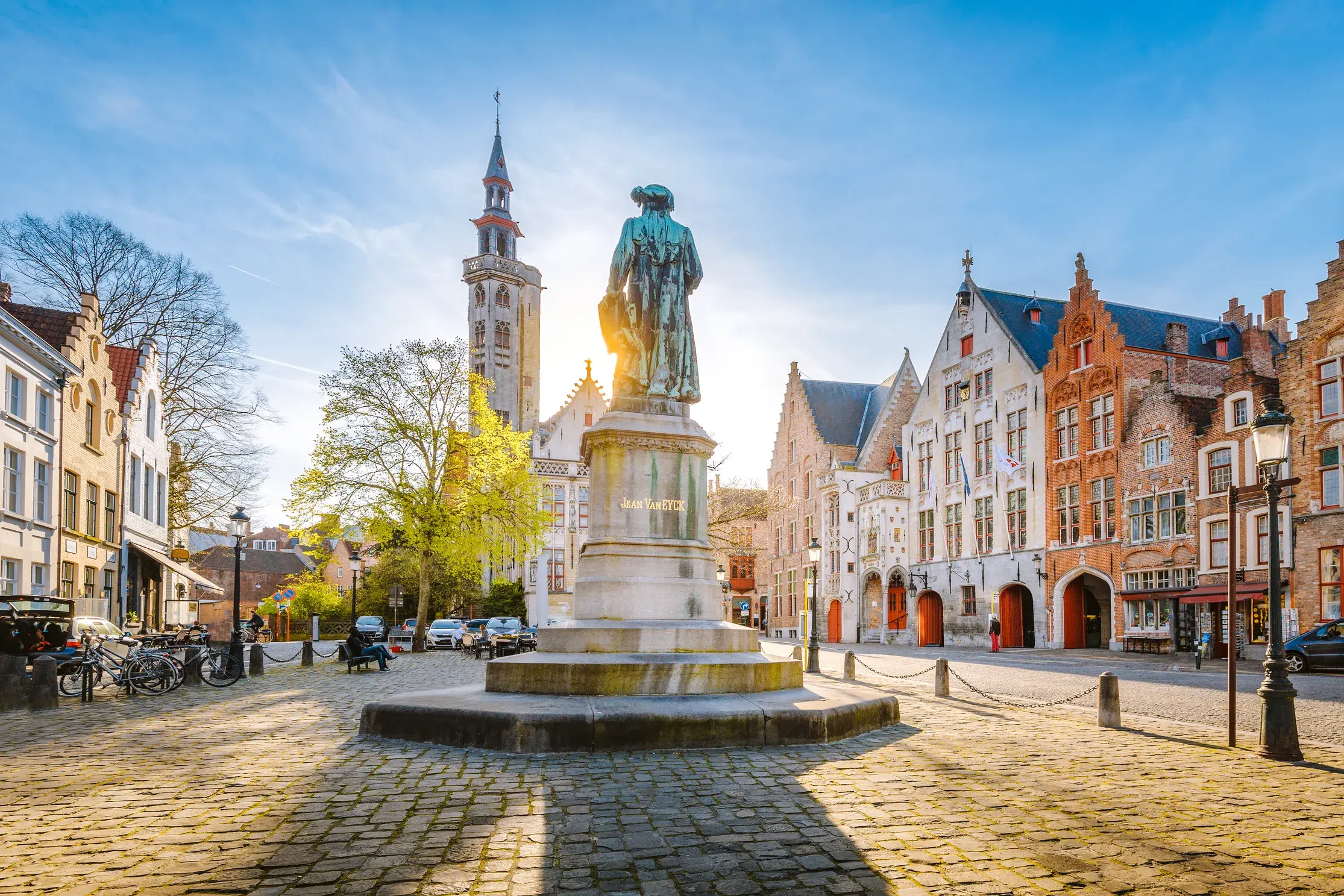 Historischer Jan-van-Eyck-Platz in Brügge mit Giebelhäusern und der Statue des Malers im goldenen Licht der Morgensonne.