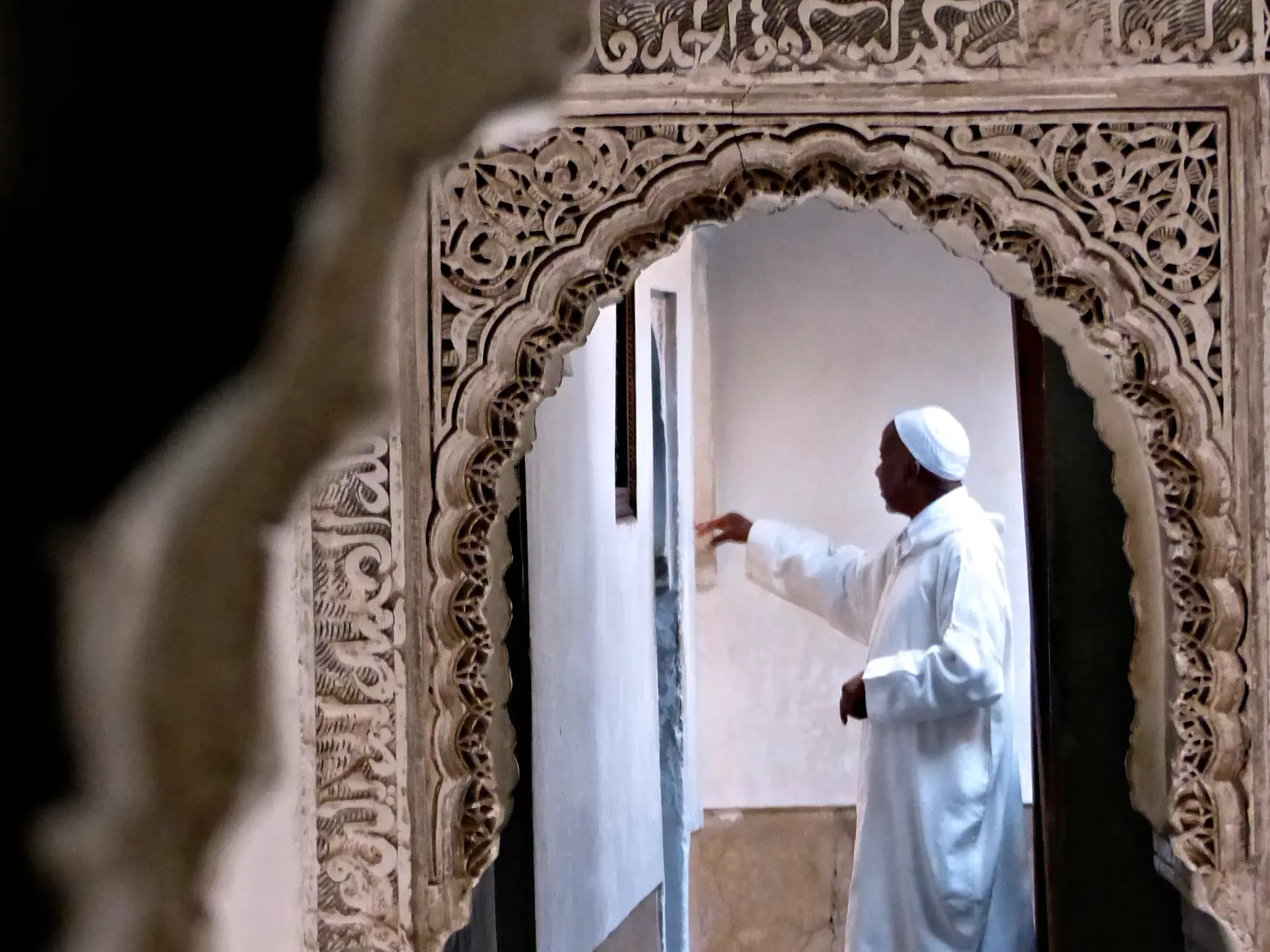 Ein Mann in traditioneller Kleidung in einem marokkanischen Riad, gesehen durch einen kunstvoll verzierten Stuckbogen.
