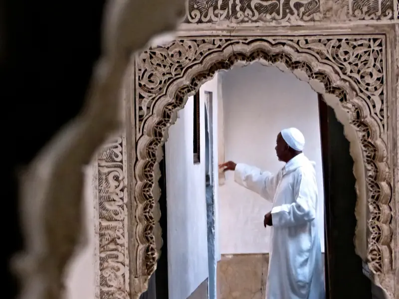 Ein Mann in traditioneller Kleidung in einem marokkanischen Riad, gesehen durch einen kunstvoll verzierten Stuckbogen.