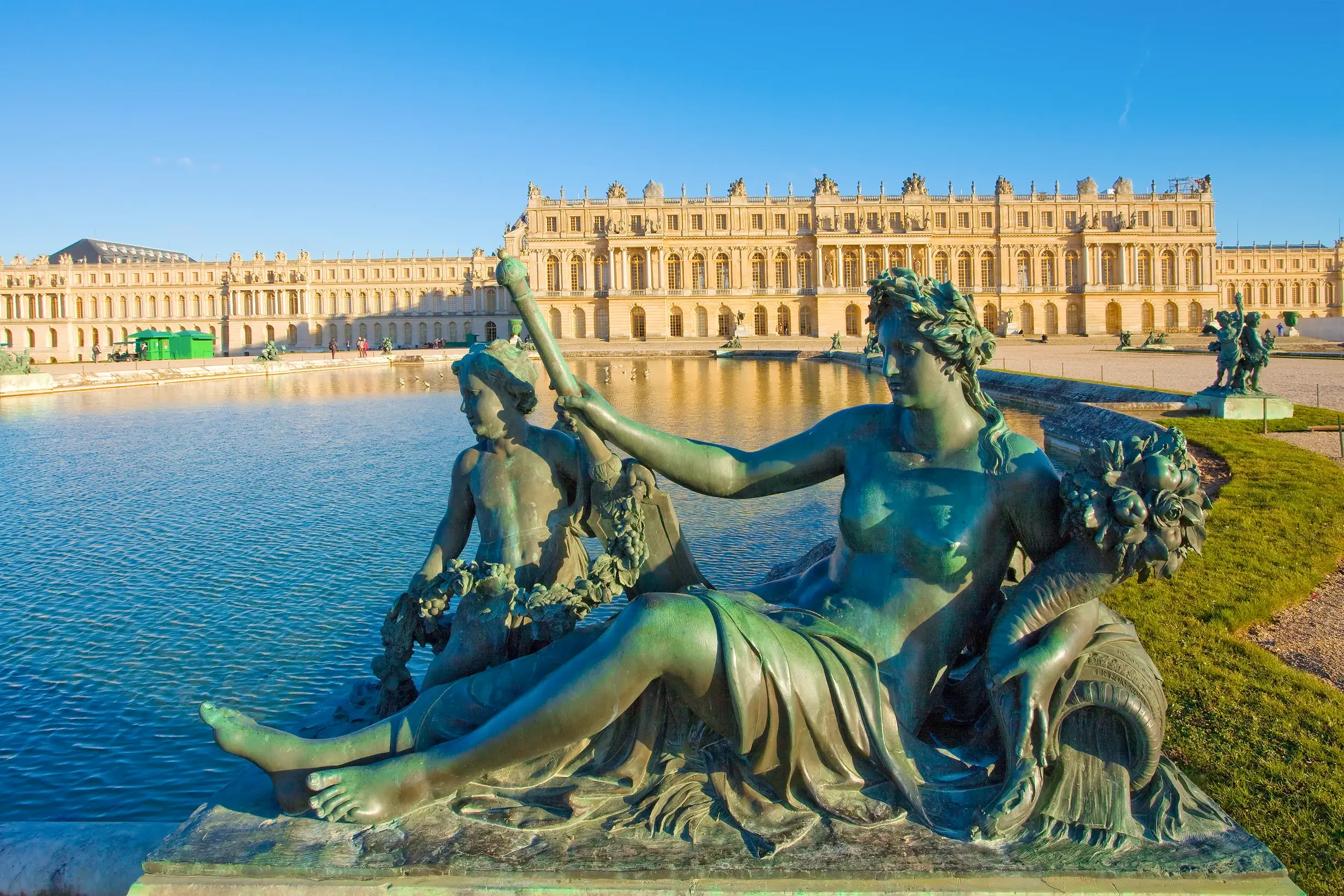 Antikisierende Bronzestatue am Rande des Parterre d'Eau mit Blick auf die sonnenbeschienene Fassade des Schlosses von Versailles.