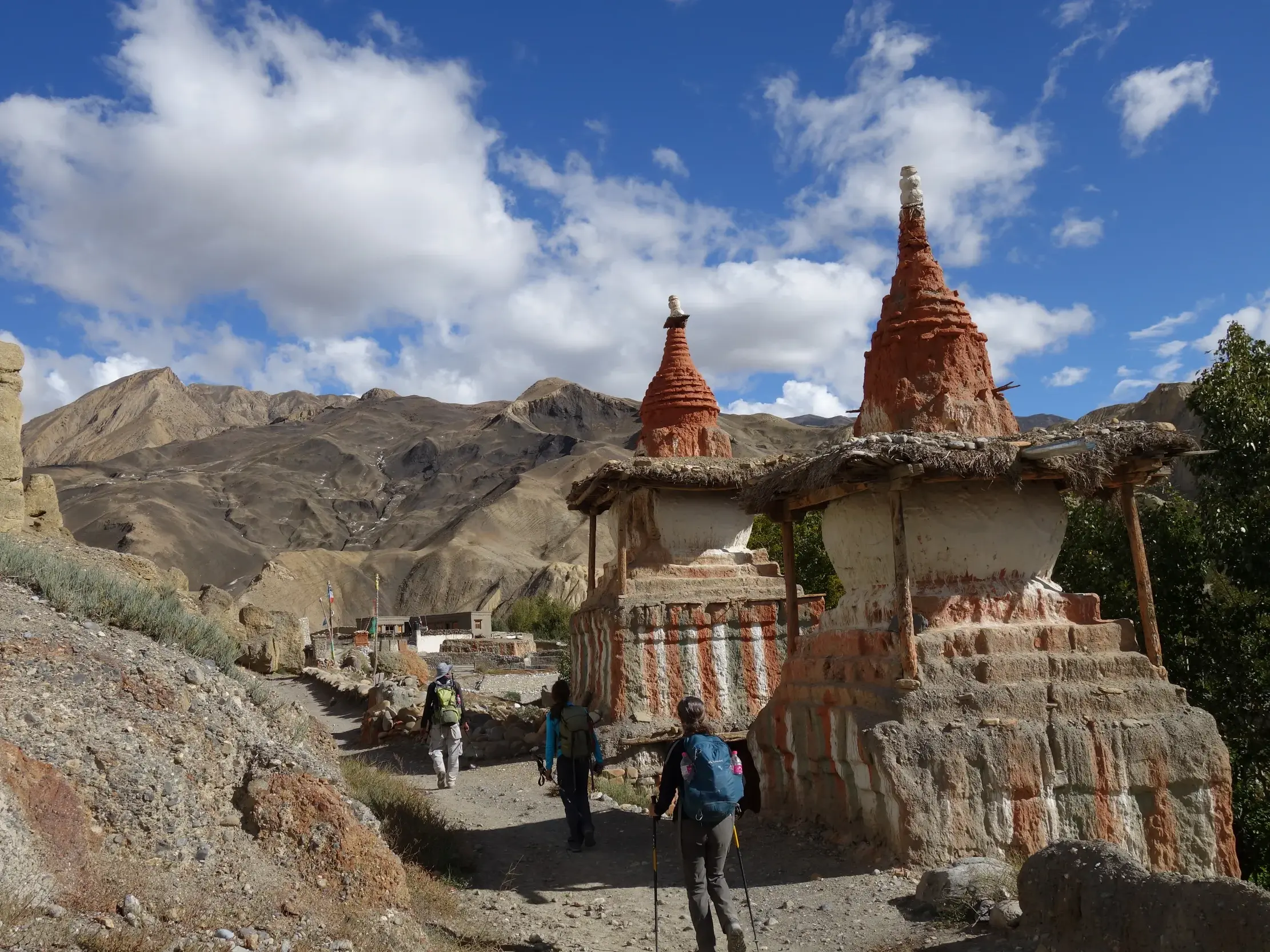 Eine kleine Trekkinggruppe wandert auf einem Pfad in der kargen Berglandschaft von Mustang vorbei an zwei ockerfarbenen Chörten.
