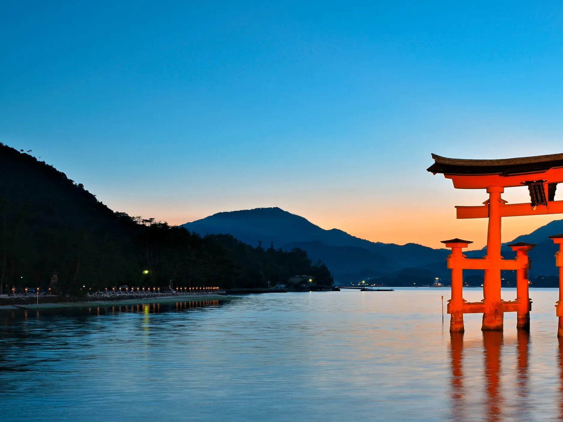 Das leuchtend rote Torii des Itsukushima-Schreins auf der Insel Miyajima bei Flut, malerisch im Wasser vor Bergen bei Dämmerung.