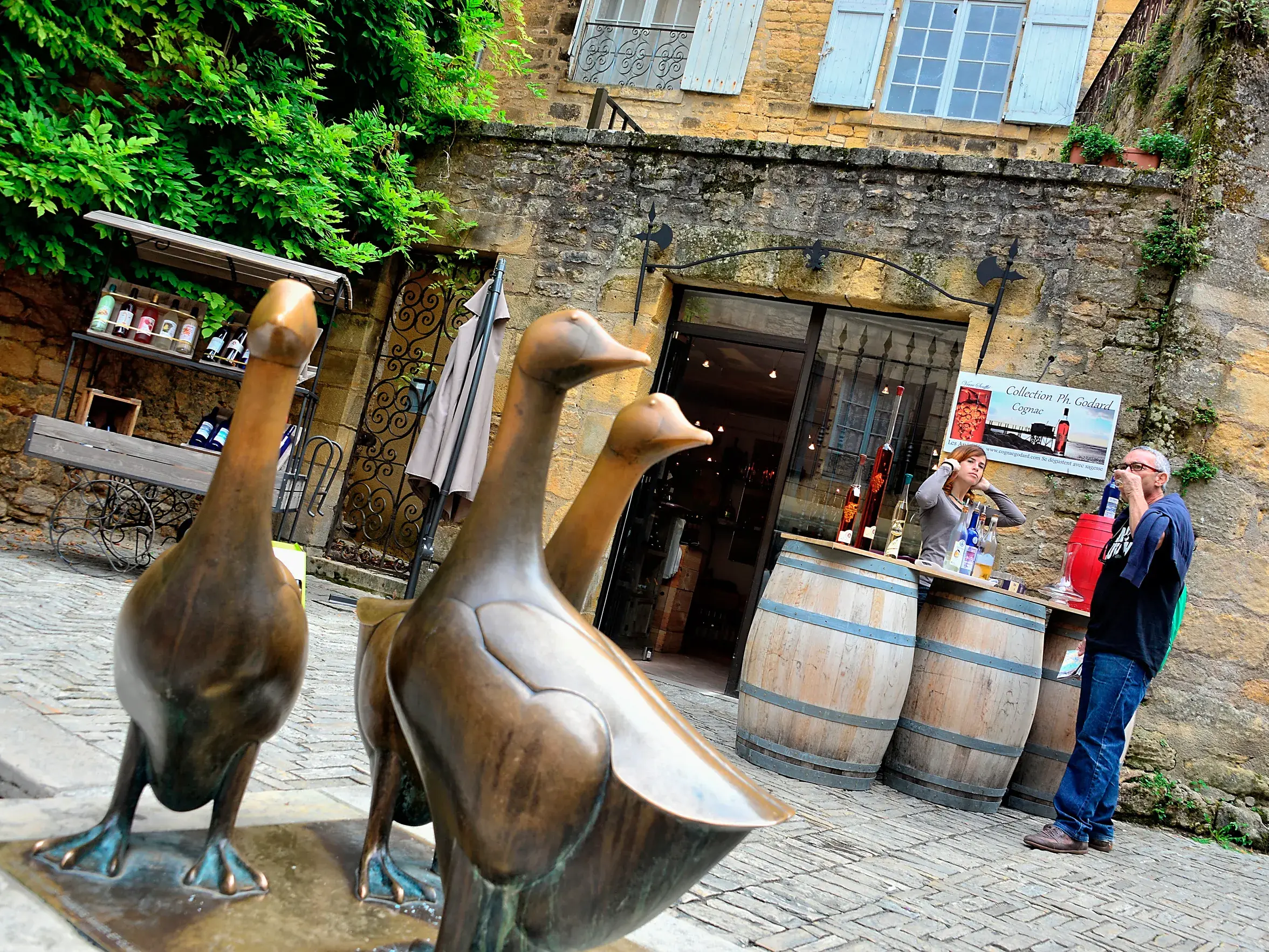 Bronzene Gänsestatuen auf einem gepflasterten Platz in Sarlat, im Hintergrund eine Verkostung an rustikalen Weinfässern.