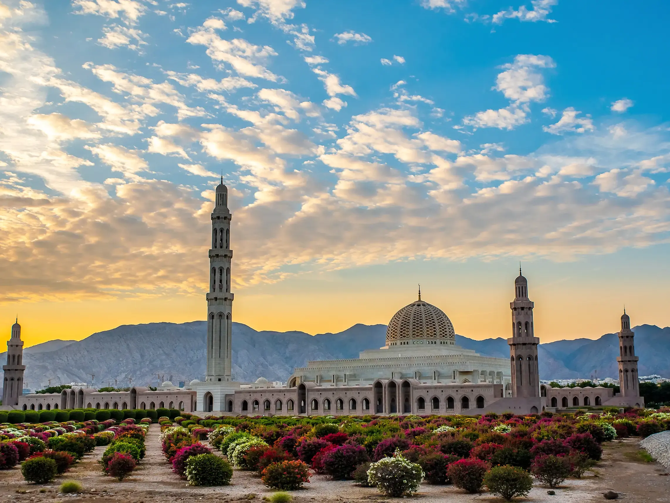 Die prachtvolle Sultan-Qaboos-Moschee in Maskat mit Gartenanlage vor der Kulisse des Hadschargebirges bei Sonnenuntergang.
