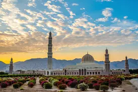 Die prachtvolle Sultan-Qaboos-Moschee in Maskat mit Gartenanlage vor der Kulisse des Hadschargebirges bei Sonnenuntergang.