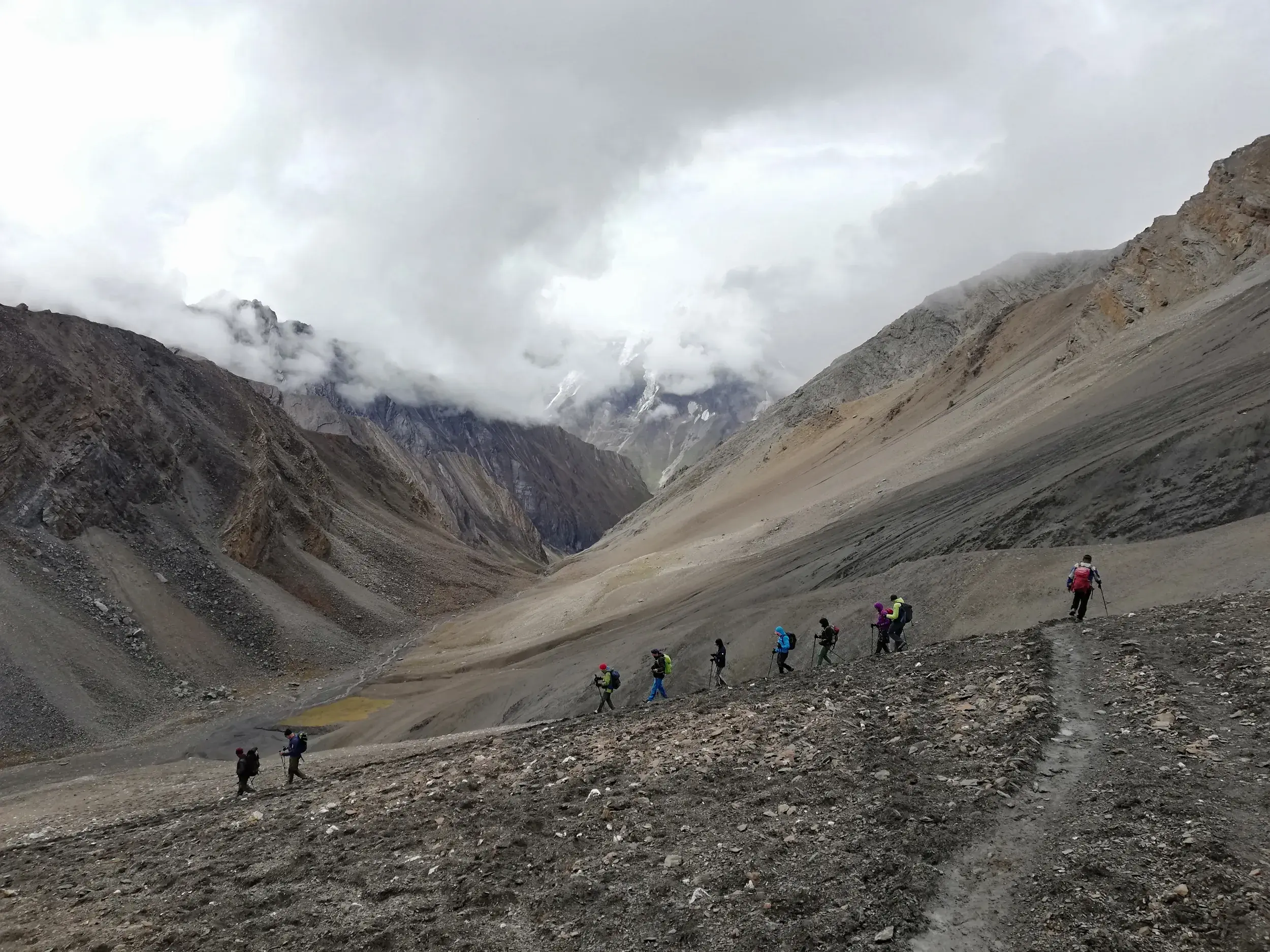Eine Trekkinggruppe quert auf einem Geröllpfad einen hohen Pass in der weiten, wolkenverhangenen Bergwelt des Inneren Dolpo.
