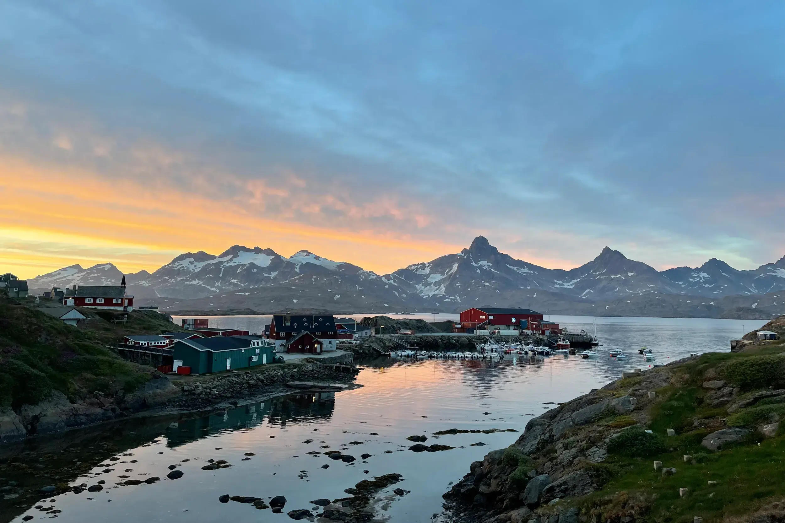Bunte Häuser von Tasiilaq am Kong-Oscar-Fjord bei Sonnenuntergang vor schneebedeckten Bergen in Ostgrönland.