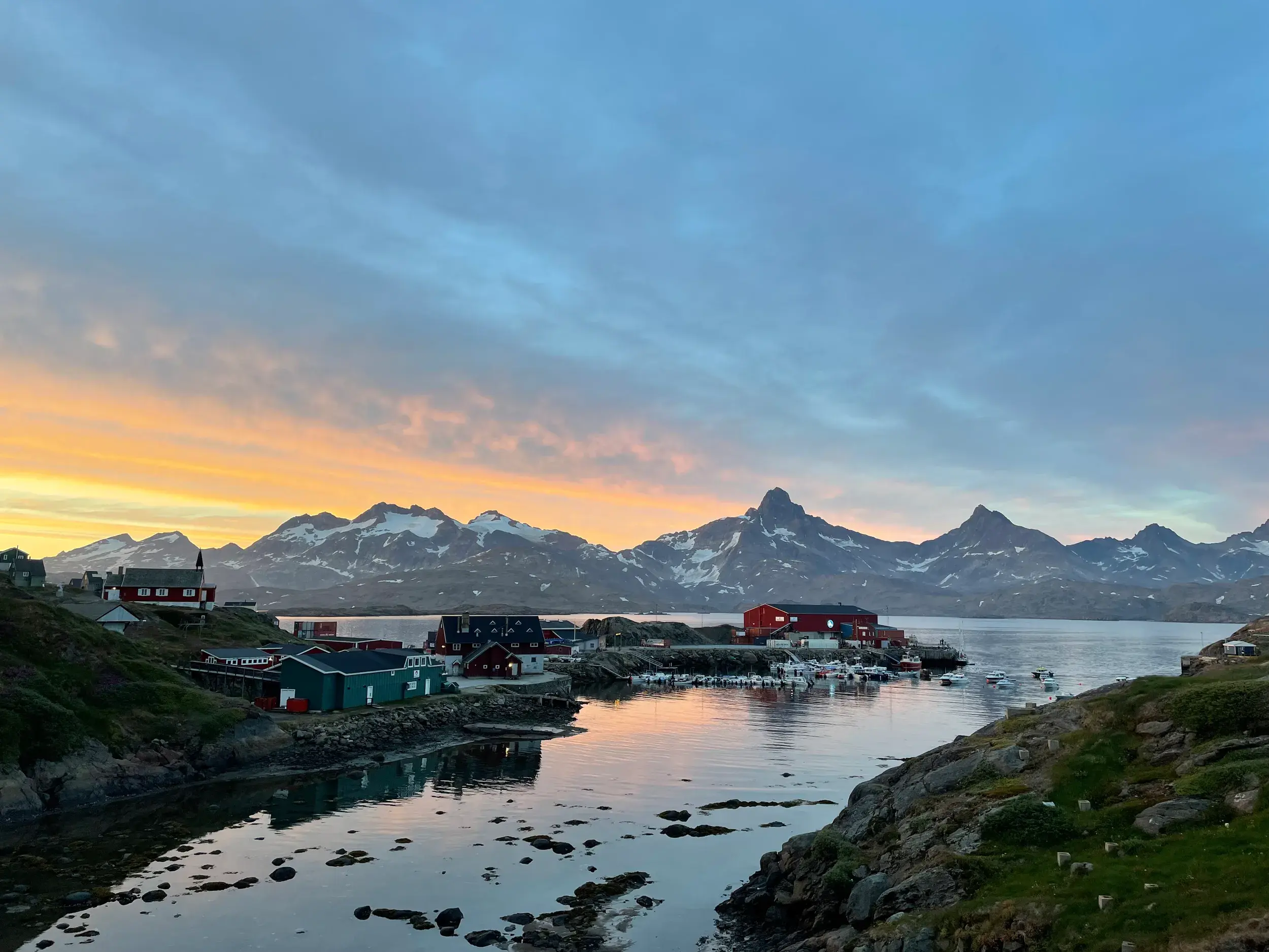Bunte Häuser von Tasiilaq am Kong-Oscar-Fjord bei Sonnenuntergang vor schneebedeckten Bergen in Ostgrönland.