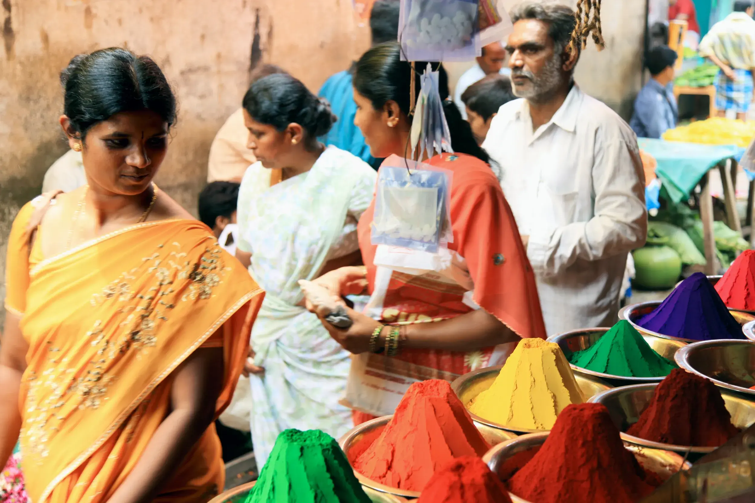 Eine Frau im Sari betrachtet leuchtend bunte Farbpulver auf einem quirligen Straßenmarkt in Madurai, Südindien.