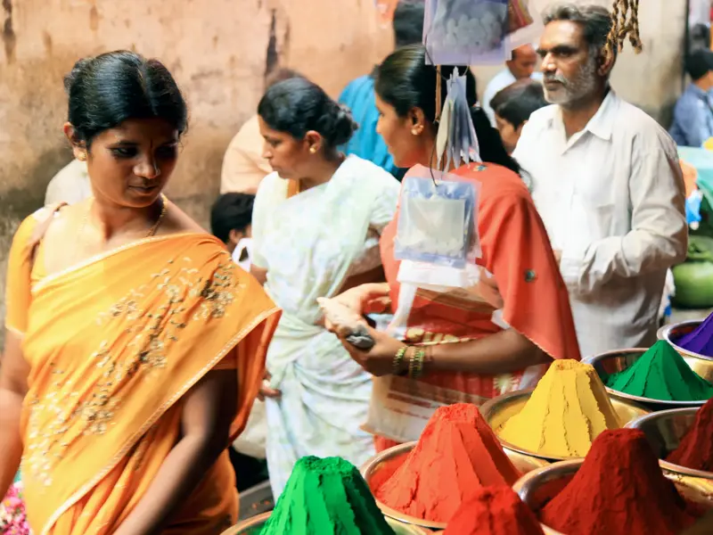 Eine Frau im Sari betrachtet leuchtend bunte Farbpulver auf einem quirligen Straßenmarkt in Madurai, Südindien.