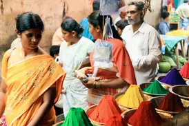Eine Frau im Sari betrachtet leuchtend bunte Farbpulver auf einem quirligen Straßenmarkt in Madurai, Südindien.