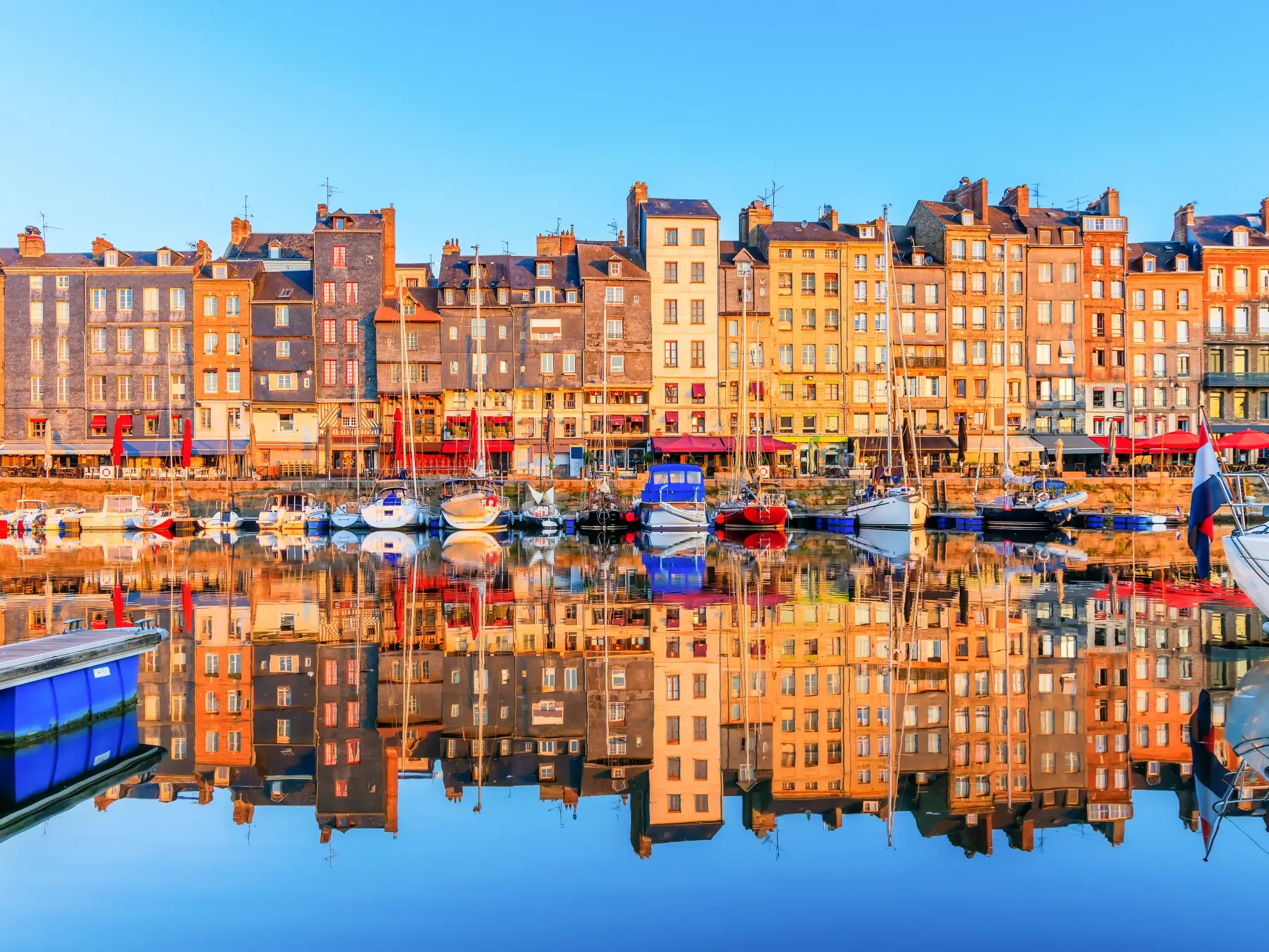 Farbenfrohe Fachwerkhäuser und Segelboote am Hafenbecken von Honfleur in der Normandie spiegeln sich im ruhigen Wasser.