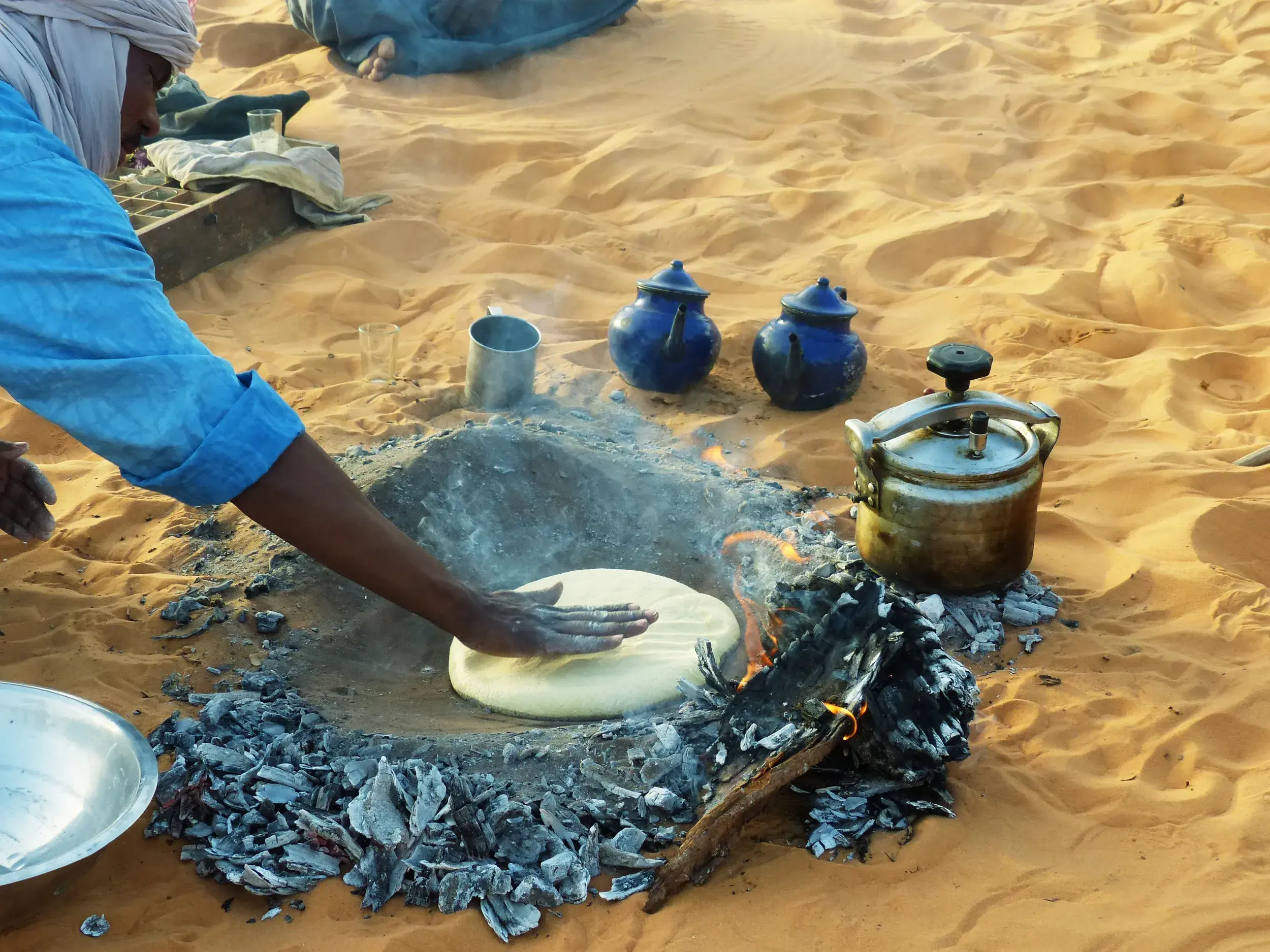 Ein Tuareg-Begleiter backt traditionelles Sandbrot in der heißen Asche eines Lagerfeuers in der Wüste Algeriens.