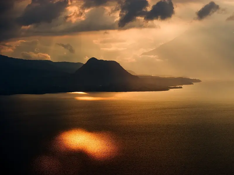 Dramatische Abendstimmung am Atitlán-See in Guatemala, goldenes Licht spiegelt sich auf dem Wasser vor Vulkansilhouetten.