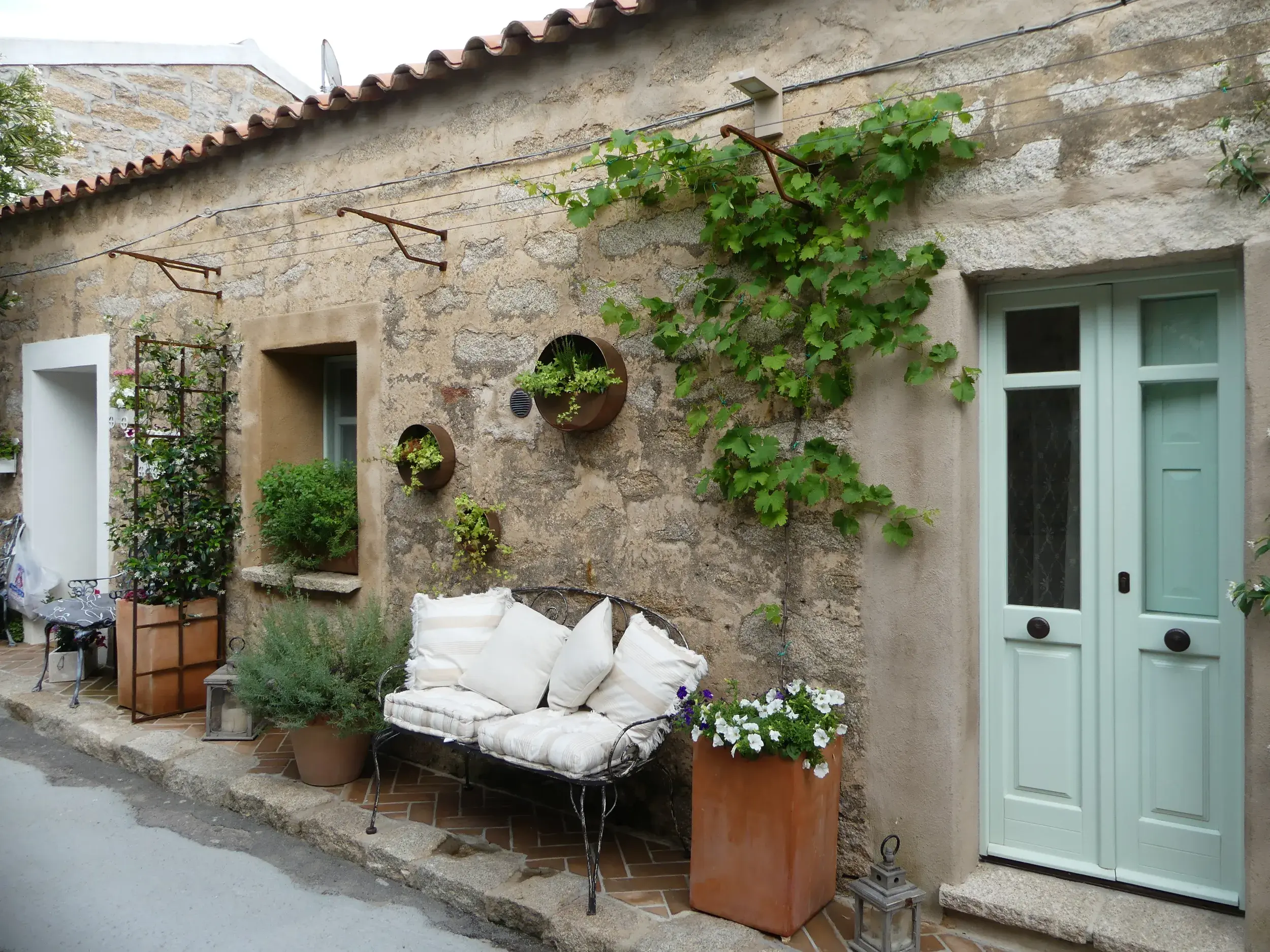 Idyllische Gasse in Santu Lussurgiu auf Sardinien mit Sitzbank vor einer rustikalen, bepflanzten Steinhausfassade.