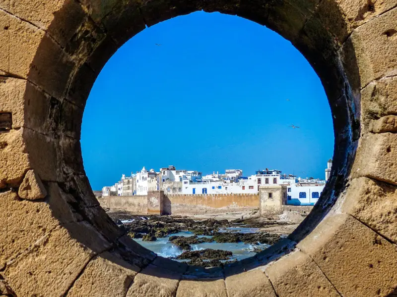 Blick durch die historische Stadtmauer auf die weiße UNESCO-Altstadt von Essaouira an der felsigen Atlantikküste Marokkos.