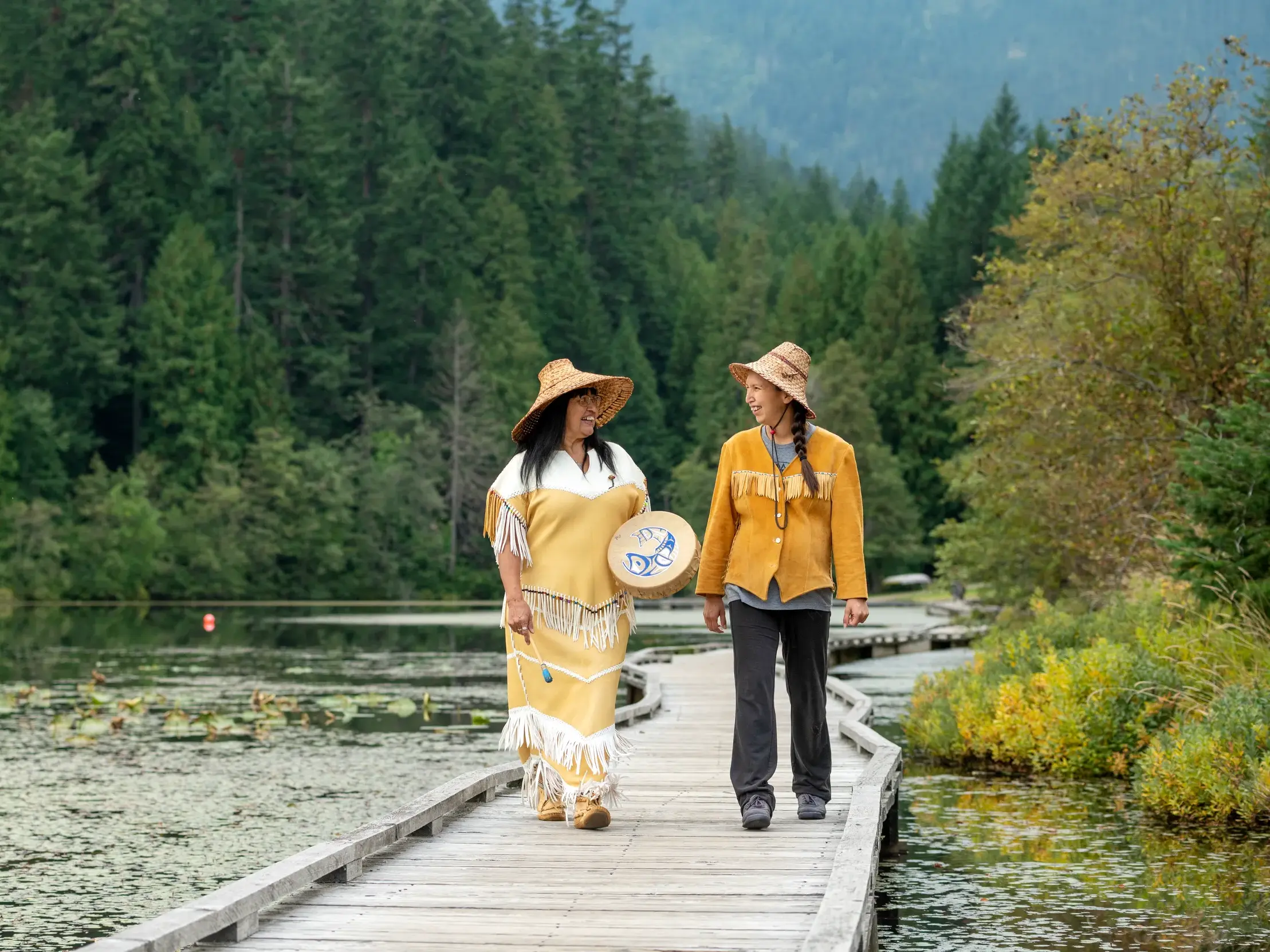 Zwei Frauen der First Nations in traditioneller Kleidung spazieren im Gespräch über einen Holzsteg an einem See in Kanada.