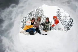 Eine fröhliche Gruppe junger Leute prostet sich mit Tassen zu, fotografiert aus einem Iglu in der verschneiten Waldlandschaft Finnlands.