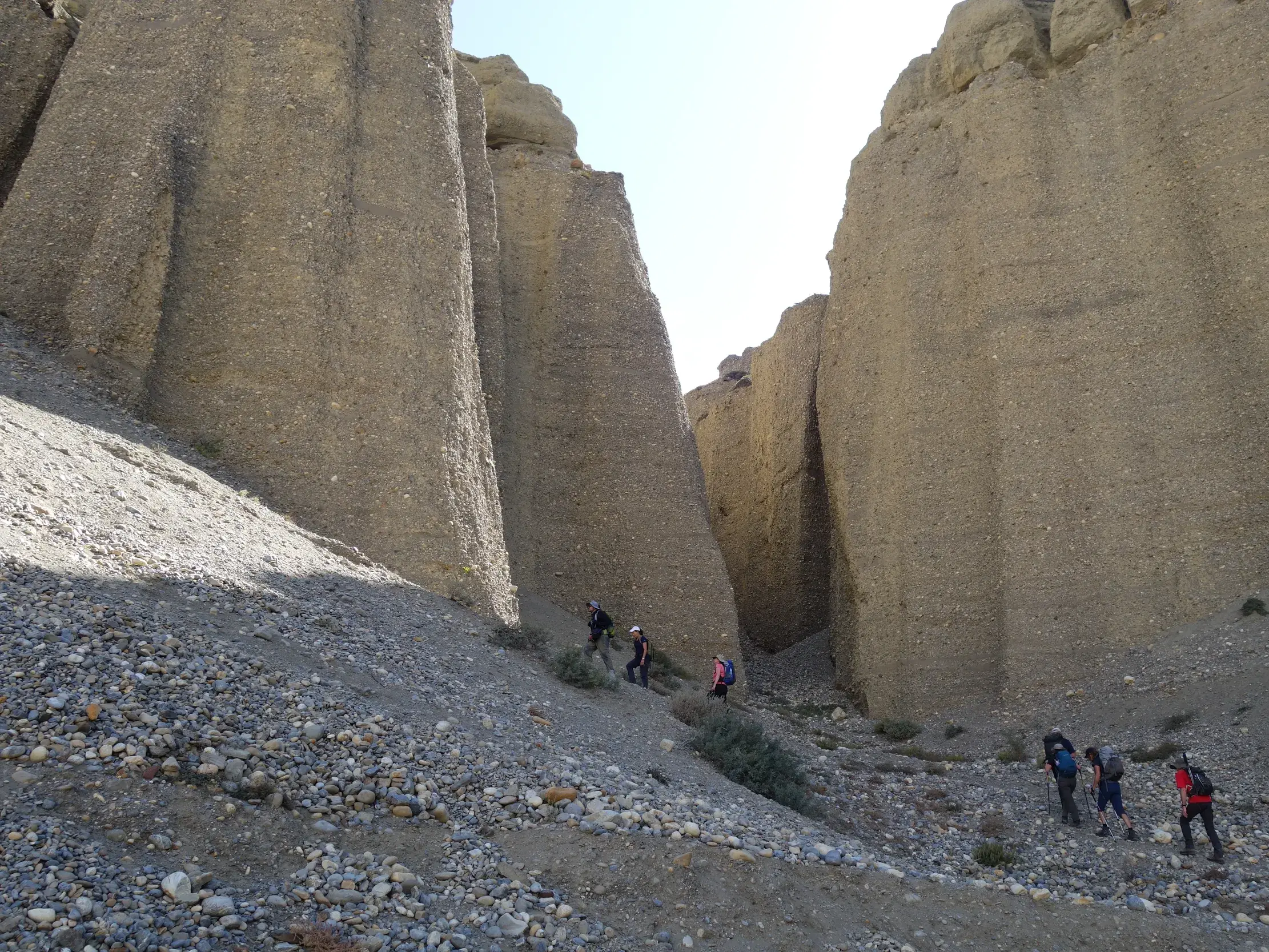 Eine kleine Wandergruppe durchquert eine beeindruckende Schlucht mit hohen, ockerfarbenen Felswänden beim Trekking in Mustang, Nepal.