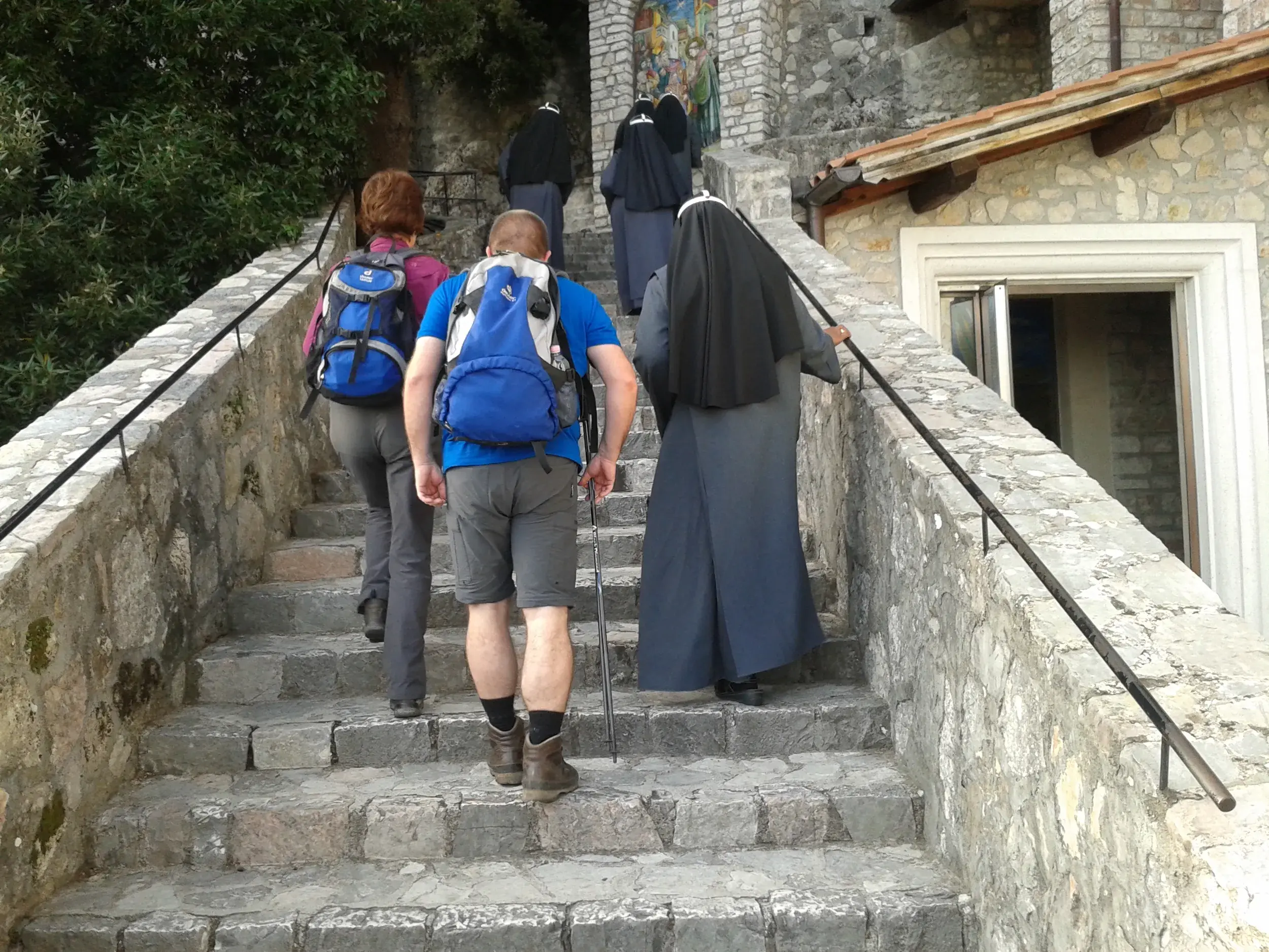 Zwei Wanderer mit Rucksäcken folgen mehreren Nonnen eine steile Steintreppe zum Kloster von Greccio in Umbrien hinauf.