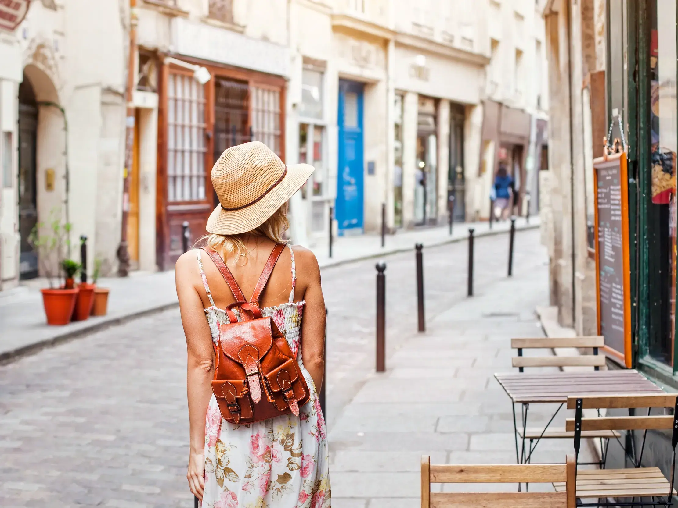 Eine Alleinreisende mit Sommerhut und Rucksack bummelt durch eine charmante Gasse in einer portugiesischen Stadt an der Algarve.
