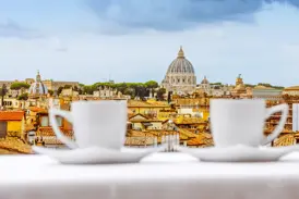 Zwei weiße Kaffeetassen auf einem Tisch mit Blick über die Dächer von Rom auf die Kuppel der Peterskirche im Vatikan.