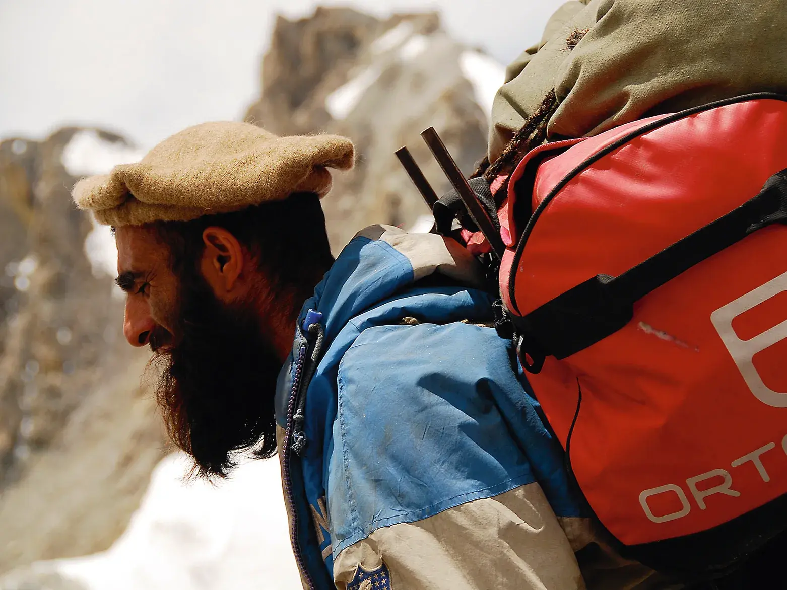 Ein einheimischer Träger mit Bart und traditioneller Mütze wandert mit einem Trekking-Rucksack durch das Karakorum-Gebirge in Pakistan.