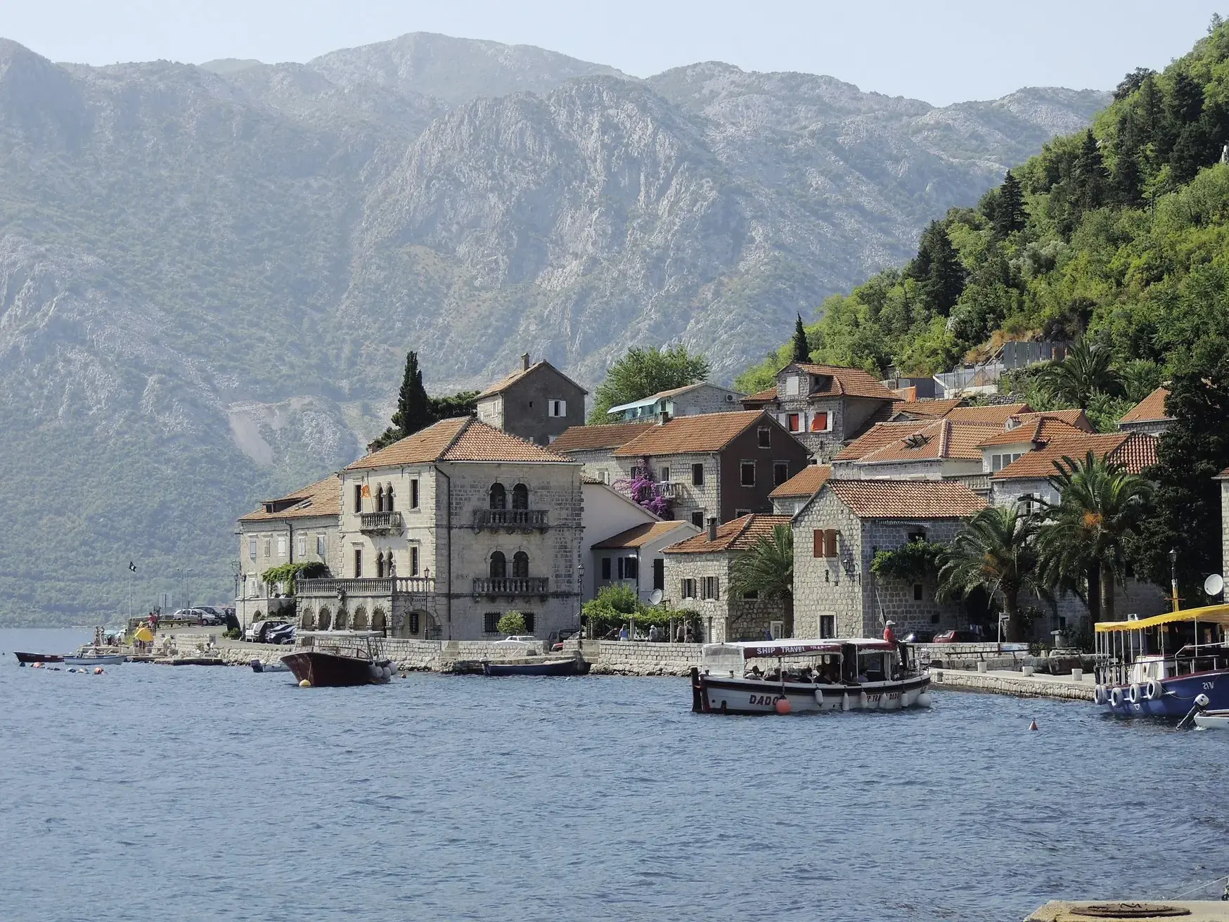 Historische Steinhäuser des Küstenortes Perast am Ufer der Bucht von Kotor in Montenegro, davor ein kleines Ausflugsboot.