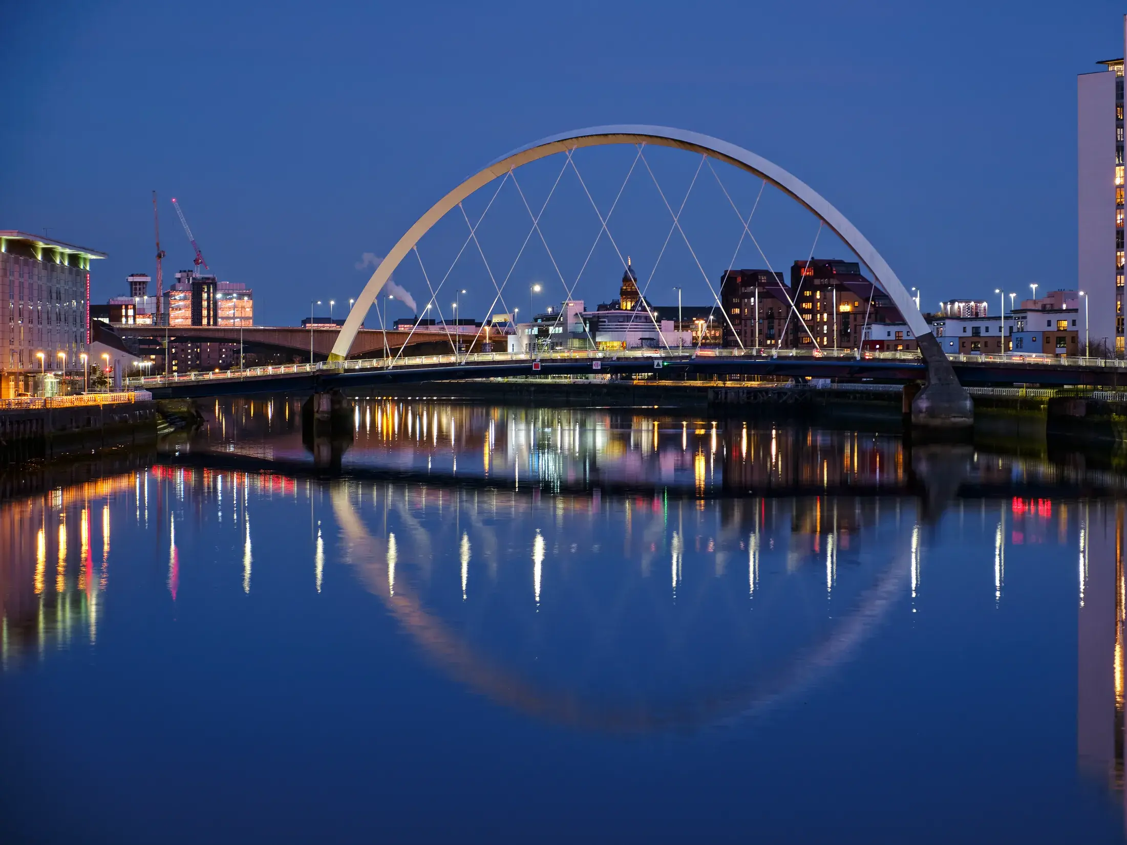 Moderne, beleuchtete Bogenbrücke an der Waterfront von Glasgow bei Nacht mit klaren Reflexionen auf dem ruhigen Wasser.