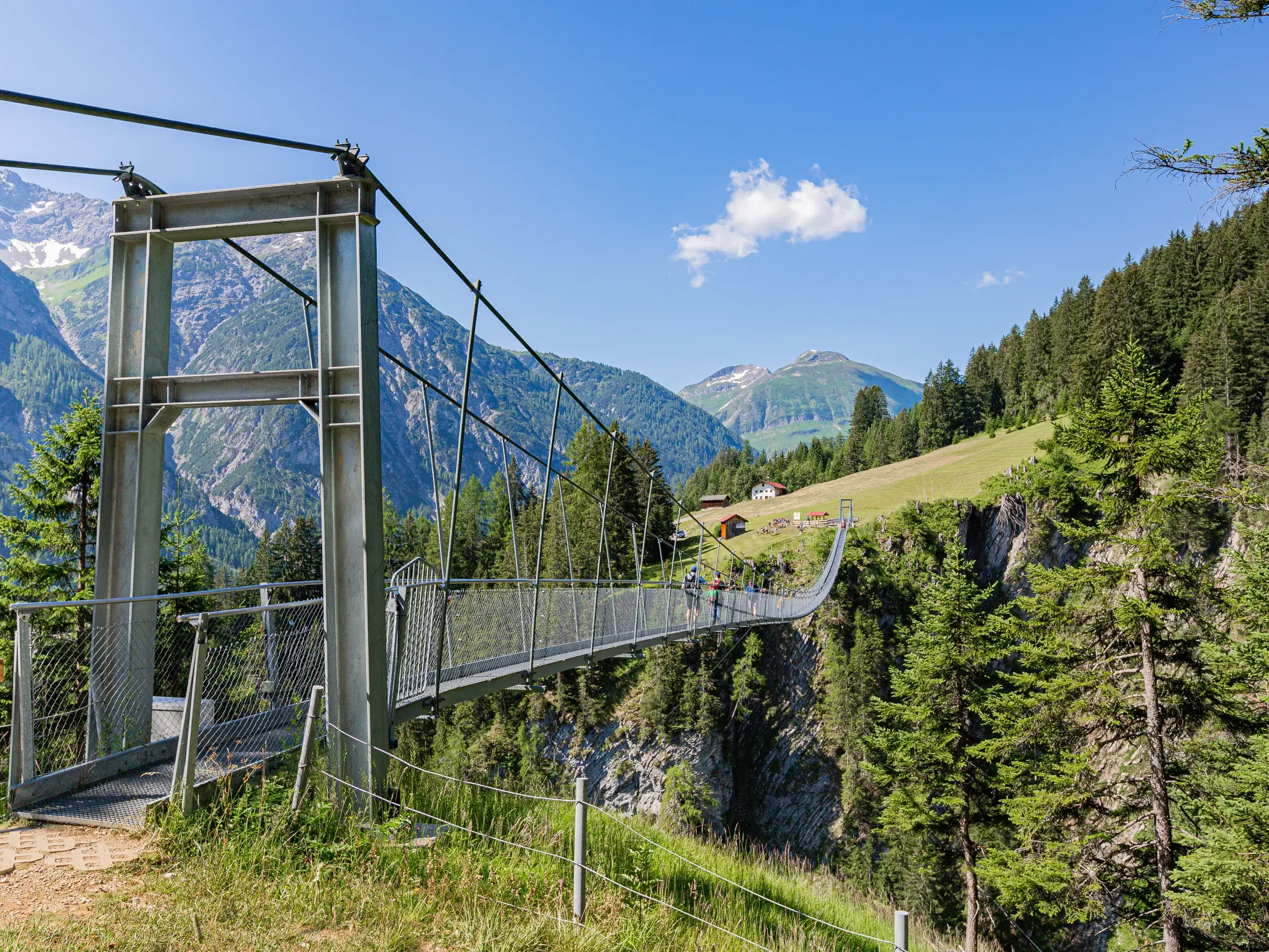 Wanderer überqueren die lange Hängebrücke Holzgau über eine tiefe Schlucht in den sonnigen Lechtaler Alpen.