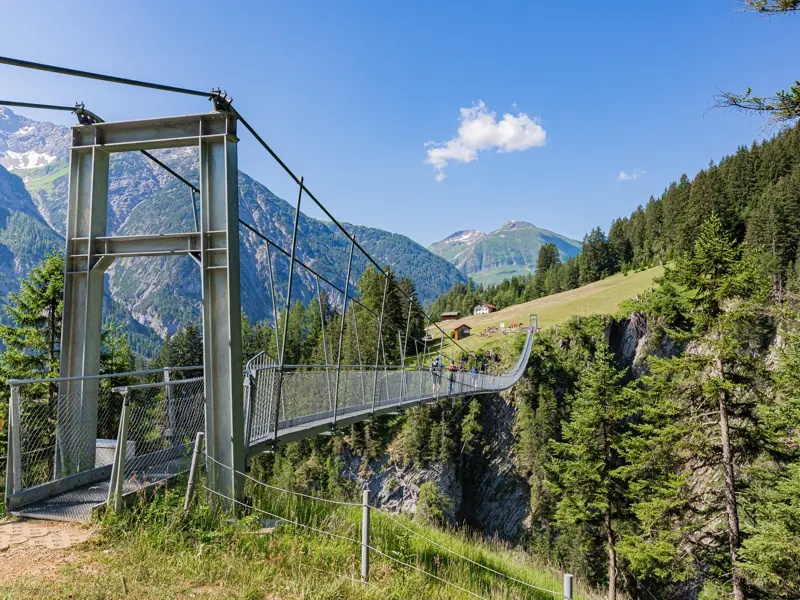 Wanderer überqueren die lange Hängebrücke Holzgau über eine tiefe Schlucht in den sonnigen Lechtaler Alpen.