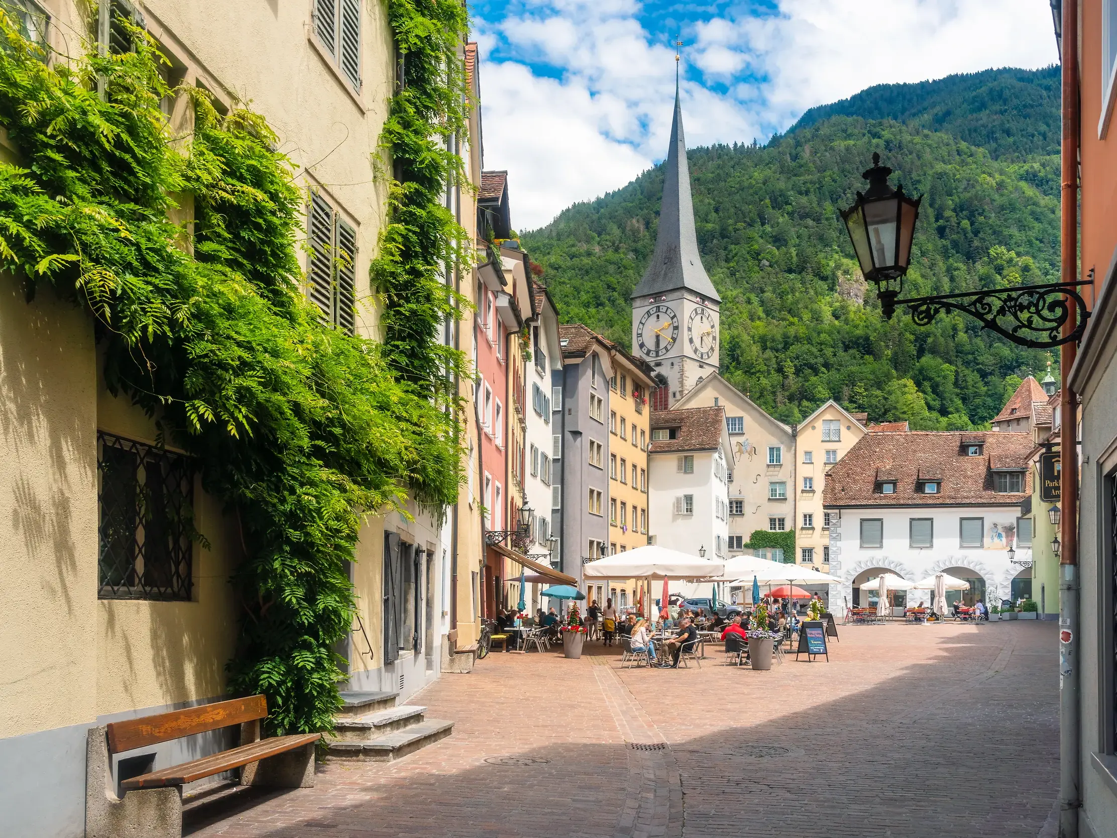 Sonnige Gasse in der Churer Altstadt mit bunten Häusern, dem Turm der Martinskirche und einem bewaldeten Berg im Hintergrund.